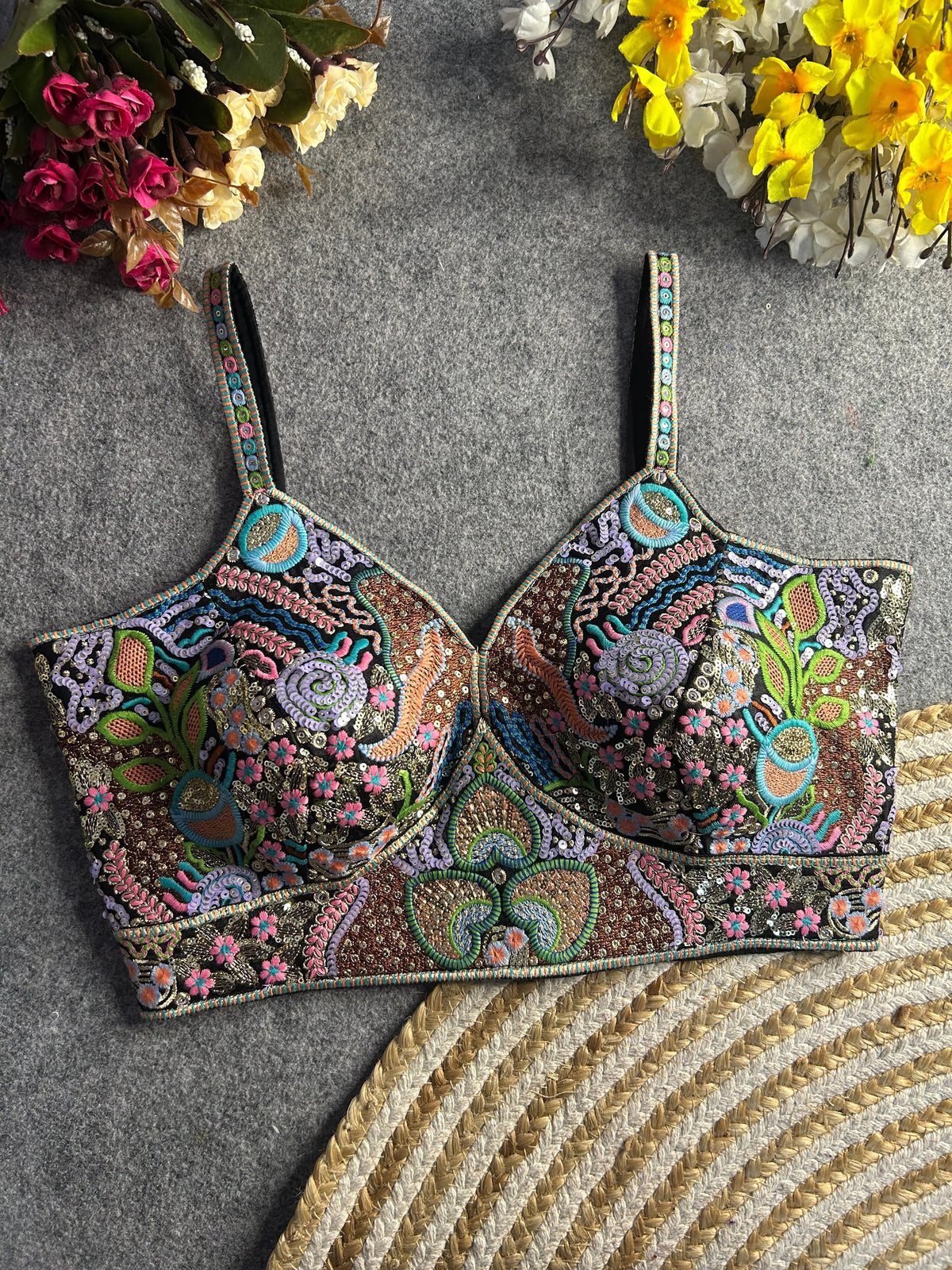 Pastel multicolor bustier-style blouse featuring intricate thread embroidery and sequin embellishments in floral, paisley, and geometric motifs, with structured cups and adjustable straps, displayed flat on a textured grey surface surrounded by artificial pink, yellow, and white flowers in the corners.