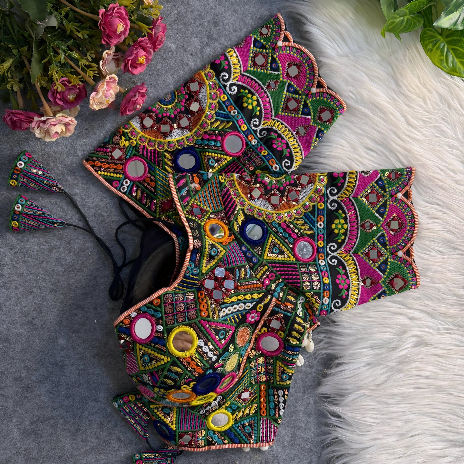 Traditional multicolor Kutch blouse featuring intricate mirror work, geometric embroidery, and vibrant threadwork in green, yellow, pink, blue, and orange. The design includes round and teardrop mirrors, beaded trim with cowrie shells, and tasseled tie-backs, displayed flat on a grey backdrop with artificial pink flowers and green foliage at the corners.