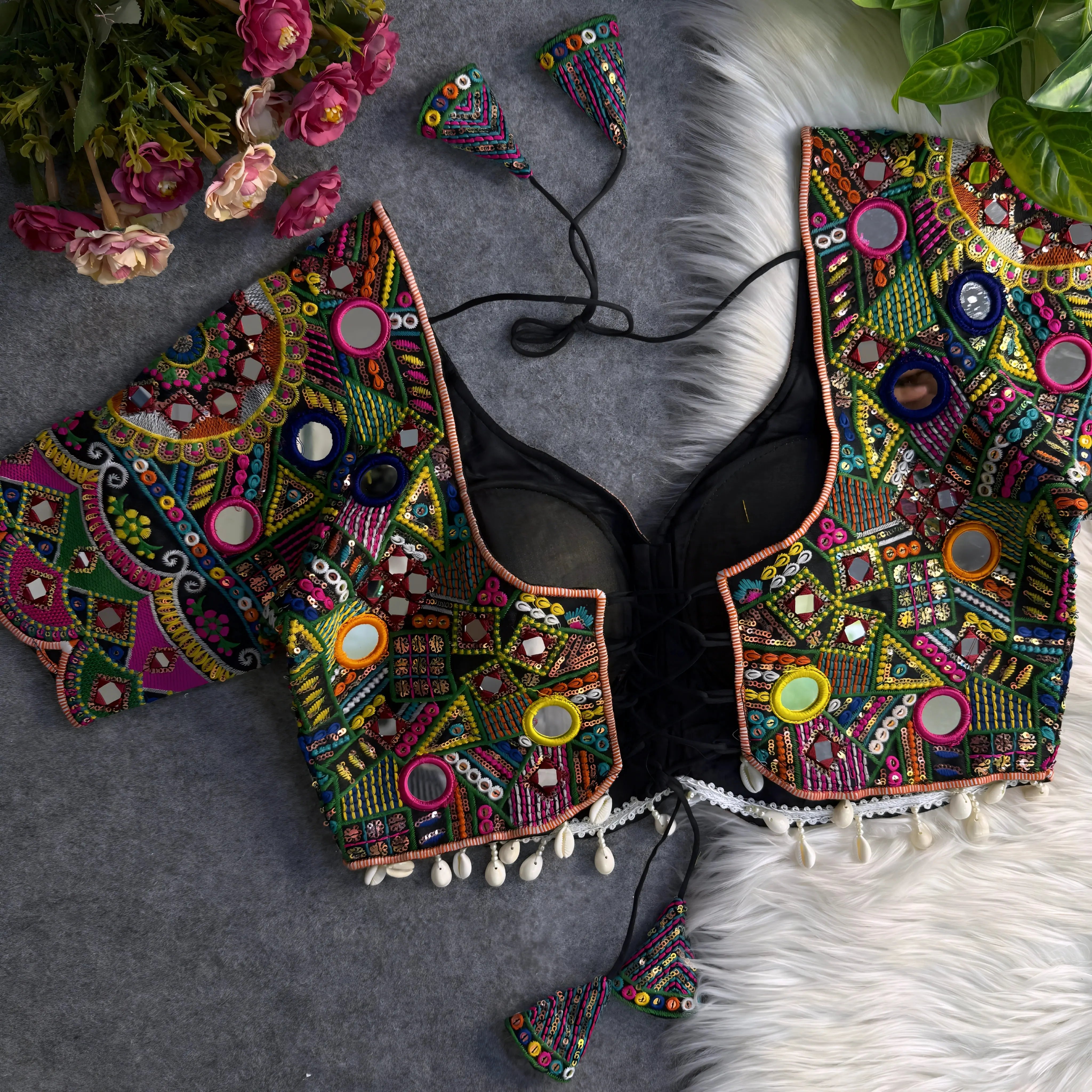 Traditional multicolor Kutch blouse featuring intricate mirror work, geometric embroidery, and vibrant threadwork in green, yellow, pink, blue, and orange. The design includes round and teardrop mirrors, beaded trim with cowrie shells, and tasseled tie-backs, displayed flat on a grey backdrop with artificial pink flowers and green foliage at the corners.