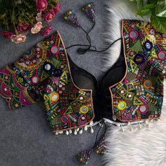 Traditional multicolor Kutch blouse featuring intricate mirror work, geometric embroidery, and vibrant threadwork in green, yellow, pink, blue, and orange. The design includes round and teardrop mirrors, beaded trim with cowrie shells, and tasseled tie-backs, displayed flat on a grey backdrop with artificial pink flowers and green foliage at the corners.