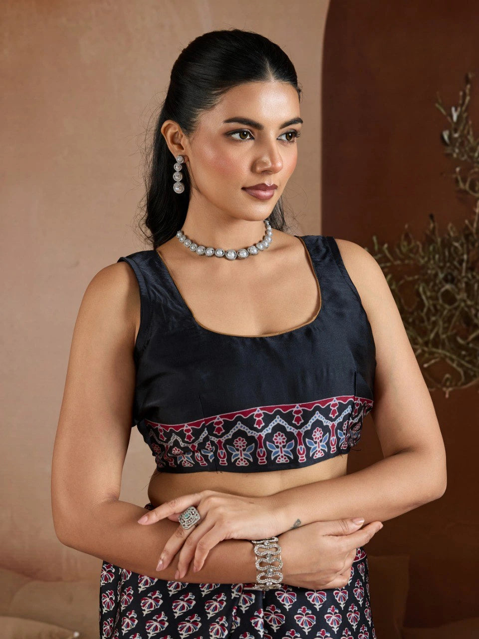 Woman wearing a black blouse with intricate patterns and jewelry against a neutral background