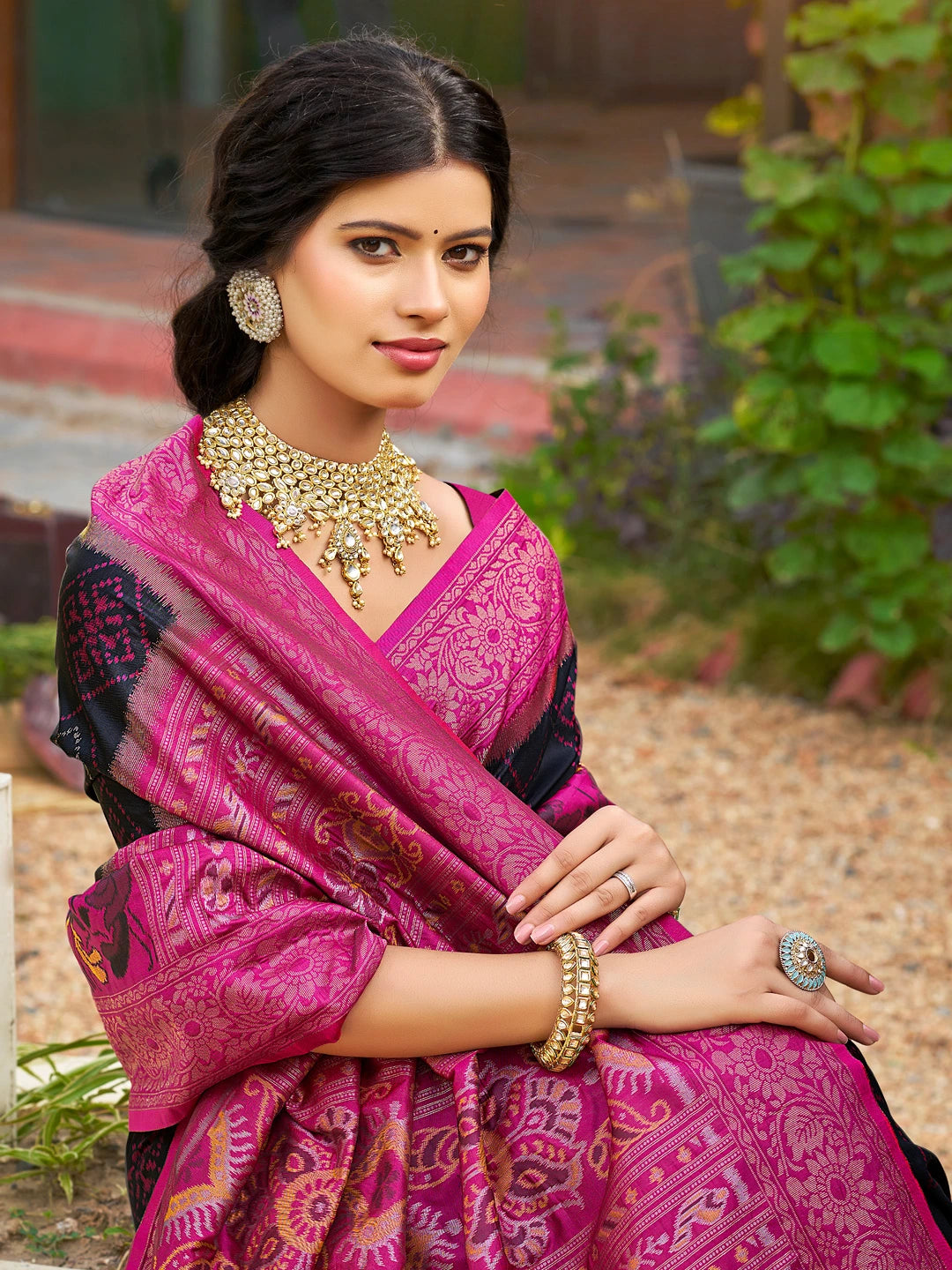Woman wearing a pink saree with intricate patterns, gold jewelry, and a blurred outdoor background.