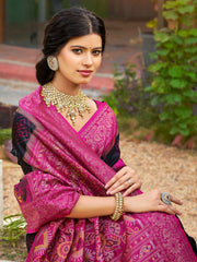 Woman wearing a pink saree with intricate patterns, gold jewelry, and a blurred outdoor background.