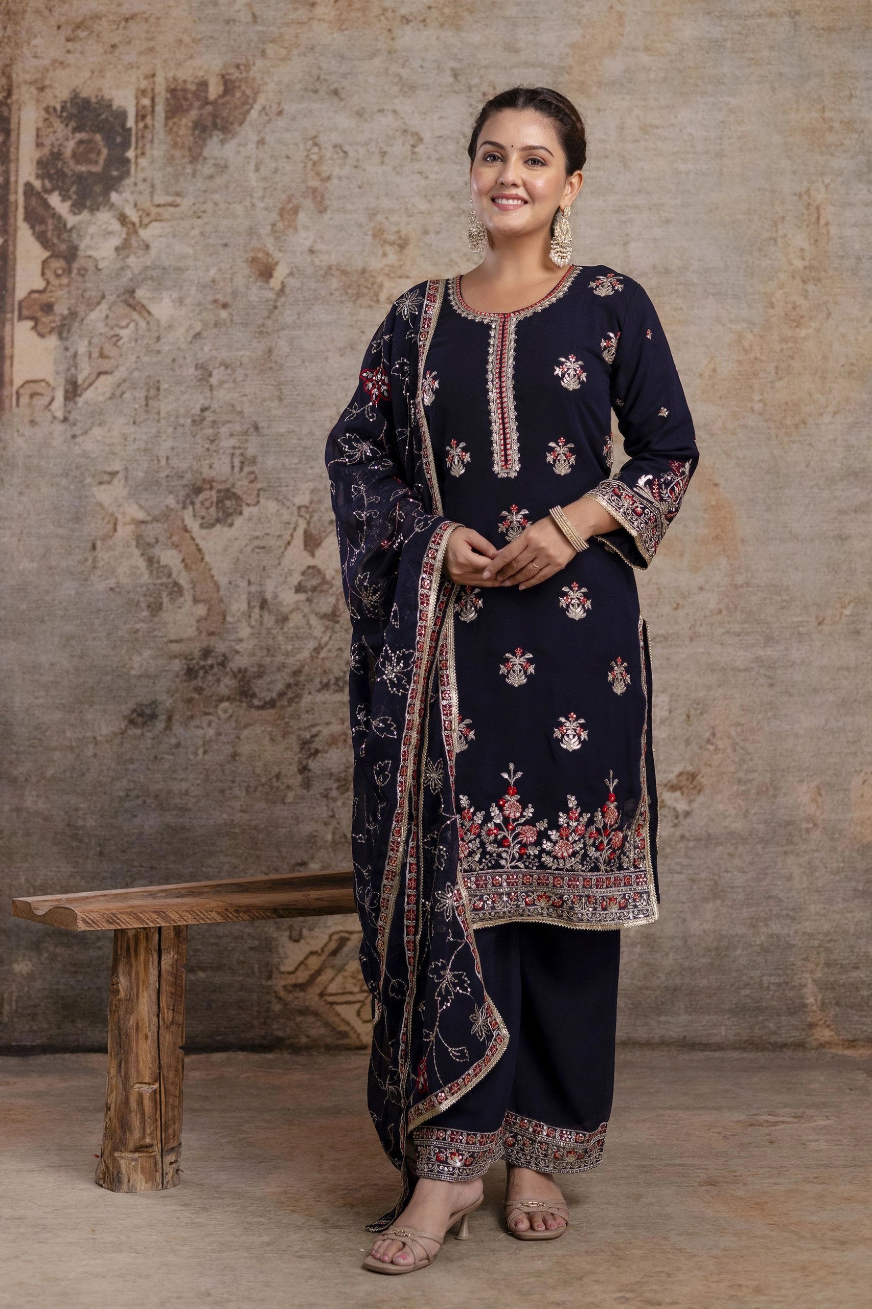 Woman wearing a dark blue embroidered traditional outfit against a textured wall.