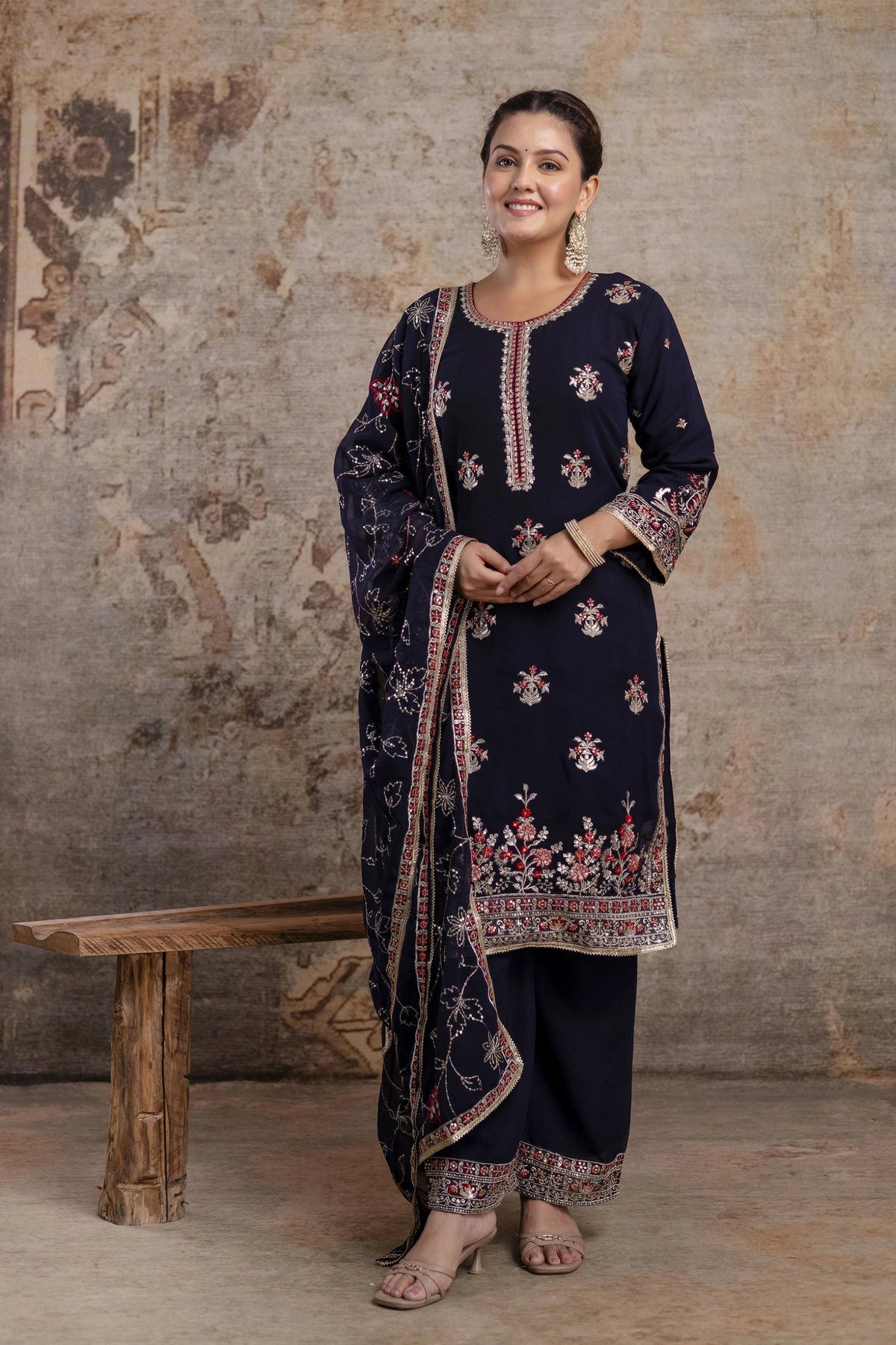 Woman wearing a dark blue embroidered traditional outfit against a textured wall.