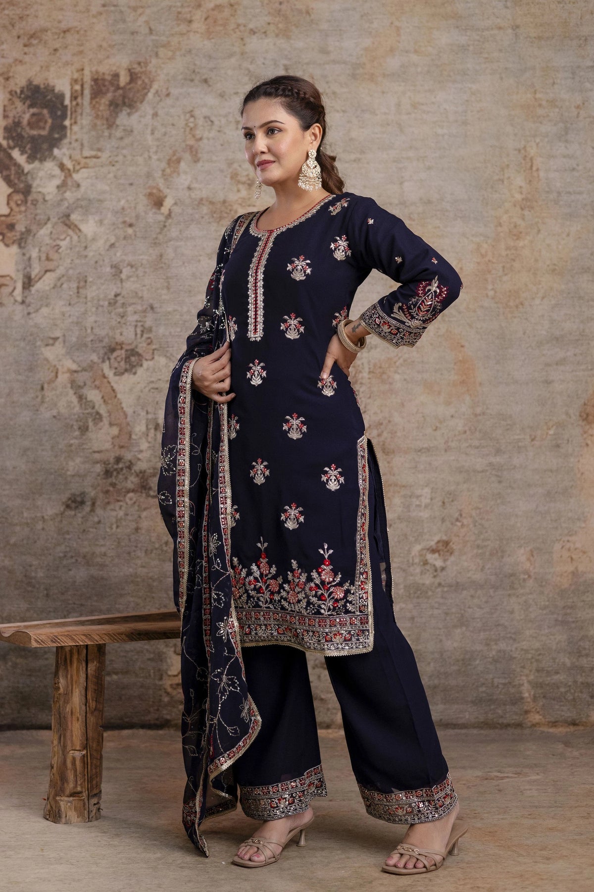 Woman wearing a navy blue embroidered traditional outfit against a textured wall.