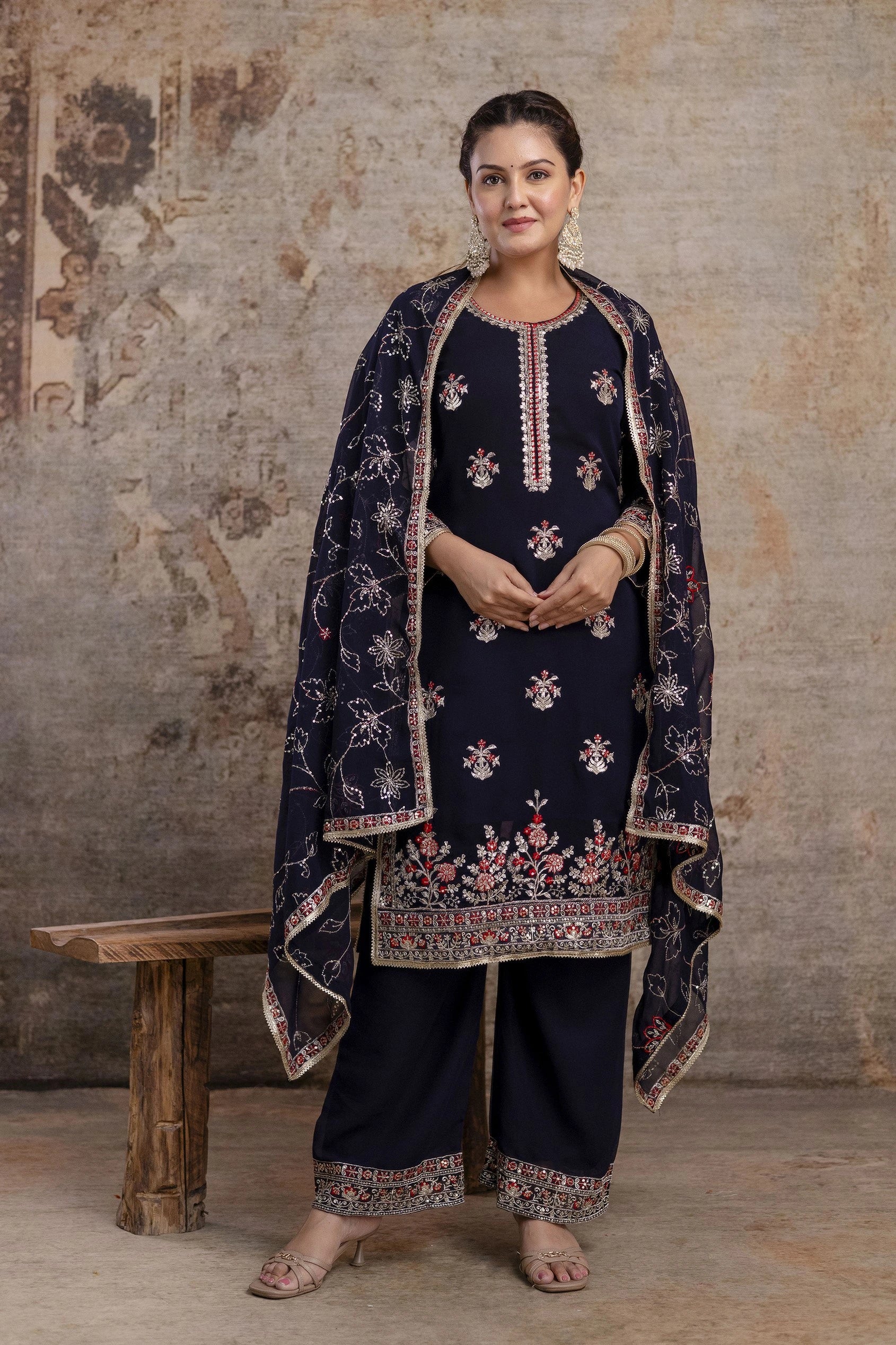 Woman wearing a traditional embroidered outfit against a textured wall.