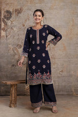Woman wearing a navy blue embroidered traditional outfit against a textured wall.