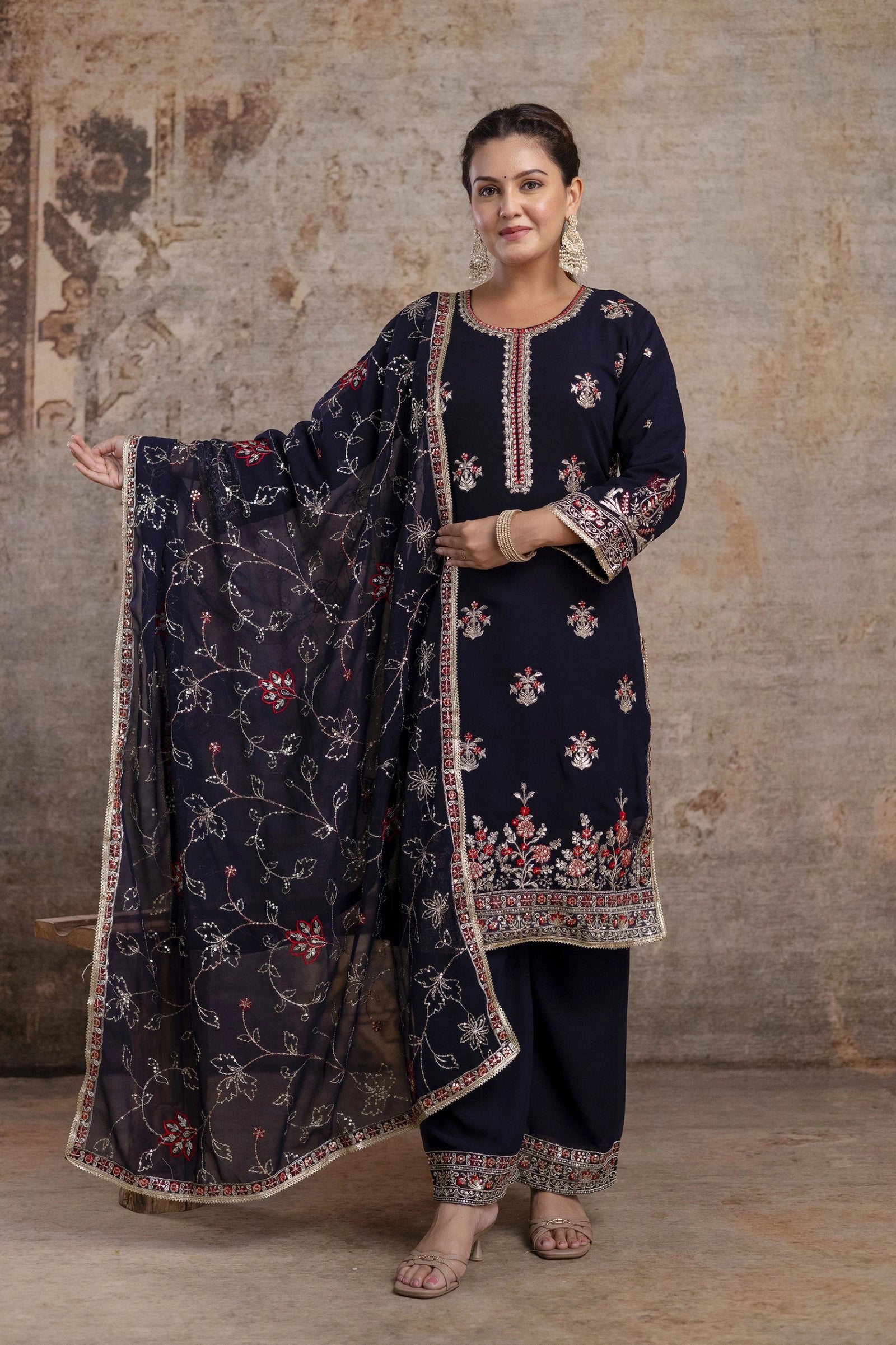 Woman wearing a navy blue embroidered traditional outfit with a matching dupatta against a textured wall.