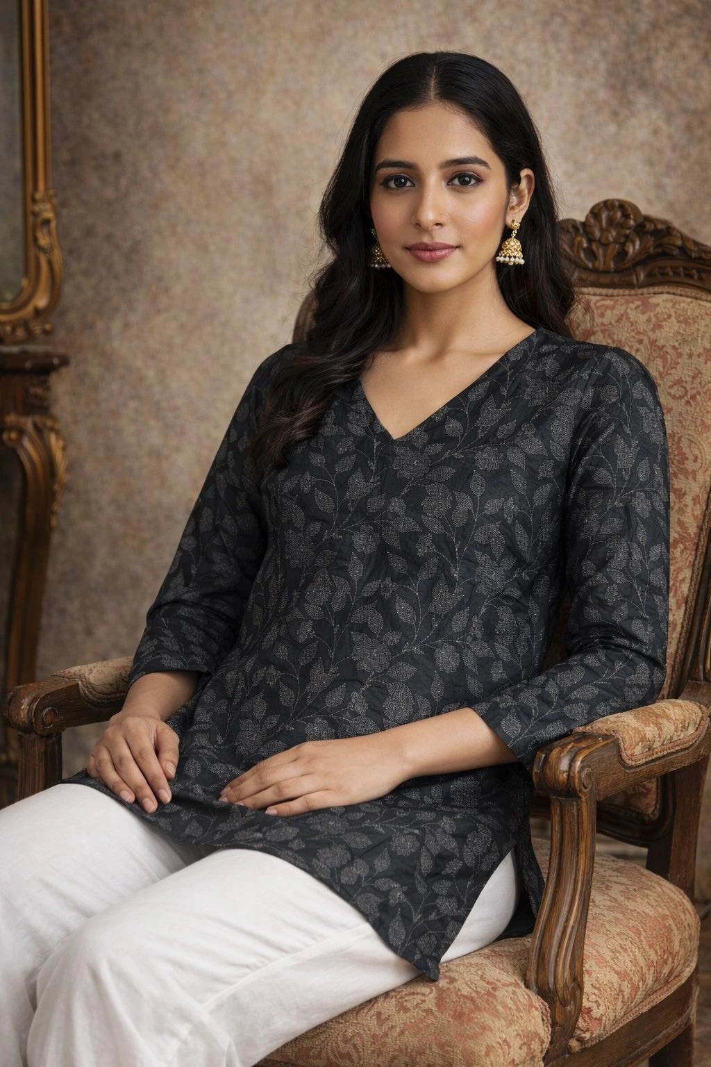 Woman sitting on a decorative chair wearing a dark patterned top and white pants.
