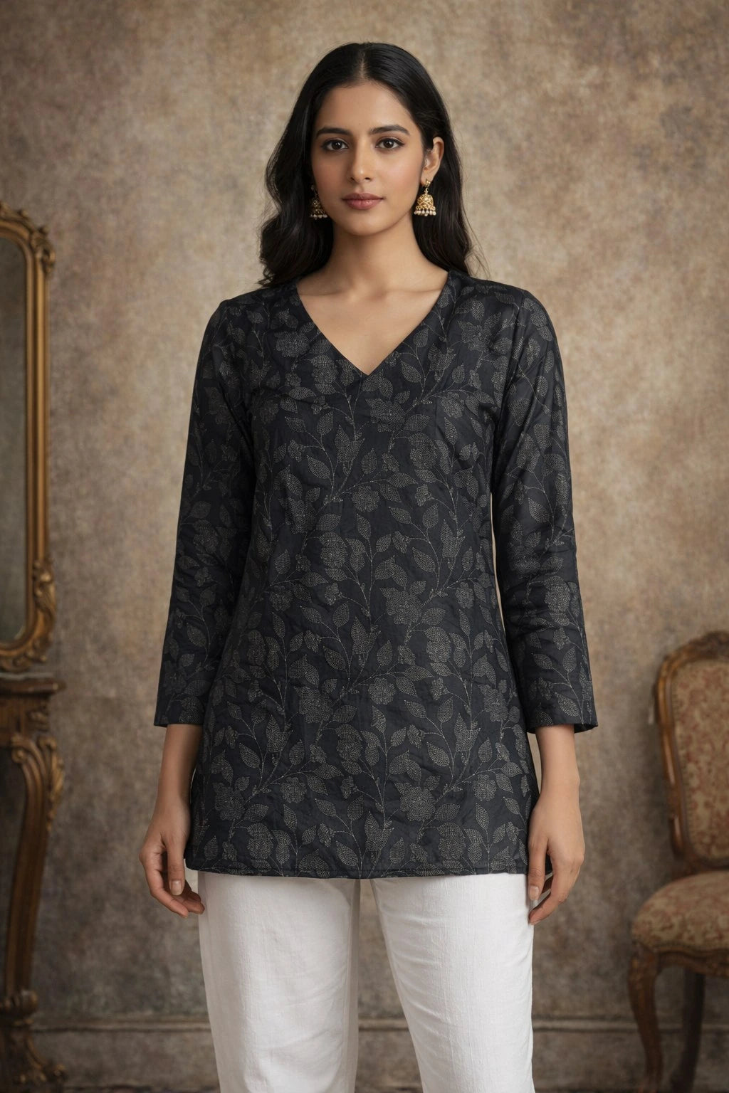 Woman wearing a dark patterned top and white pants against a textured wall.