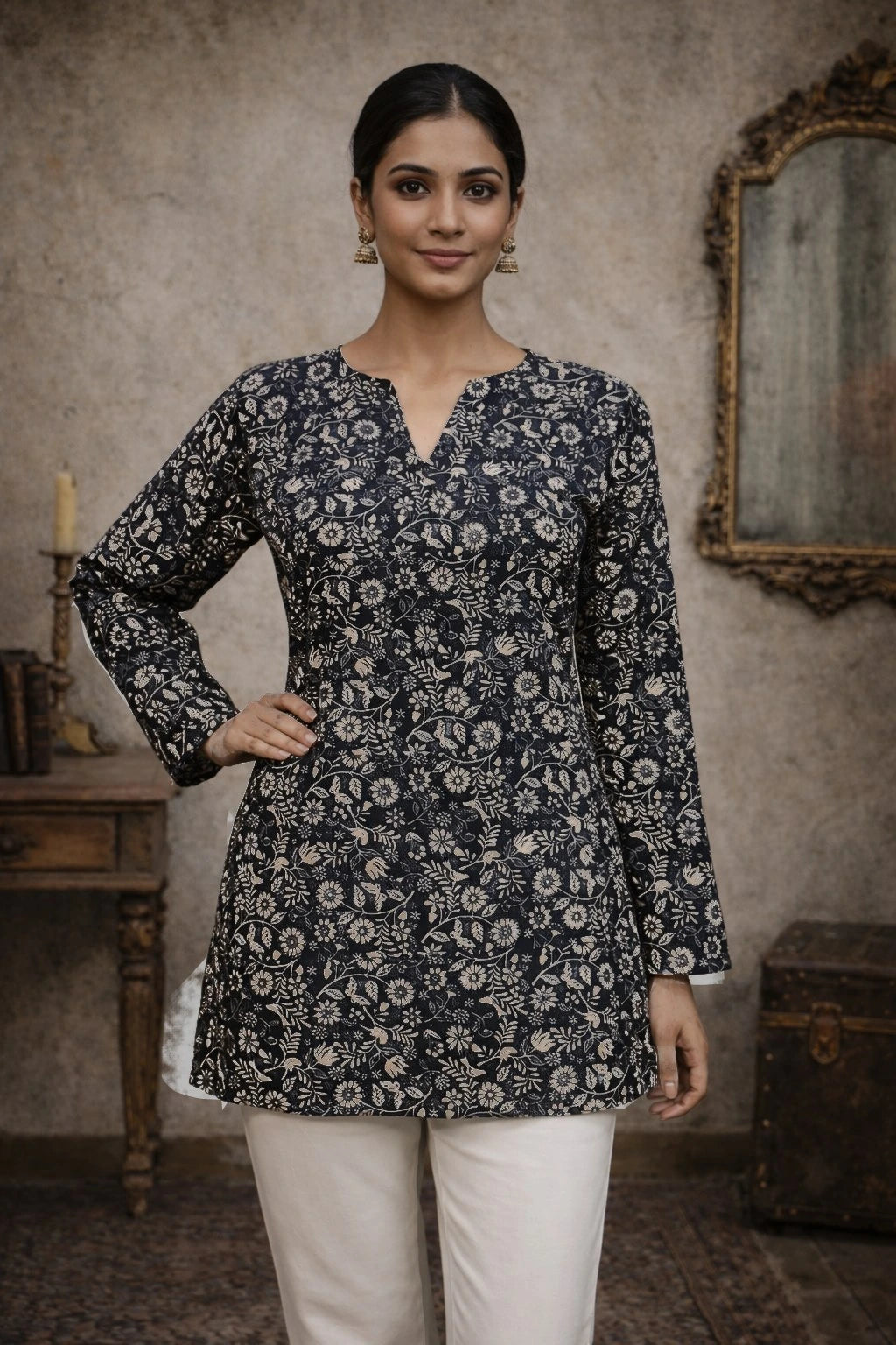 Woman wearing a dark floral patterned top with white pants in an indoor setting.
