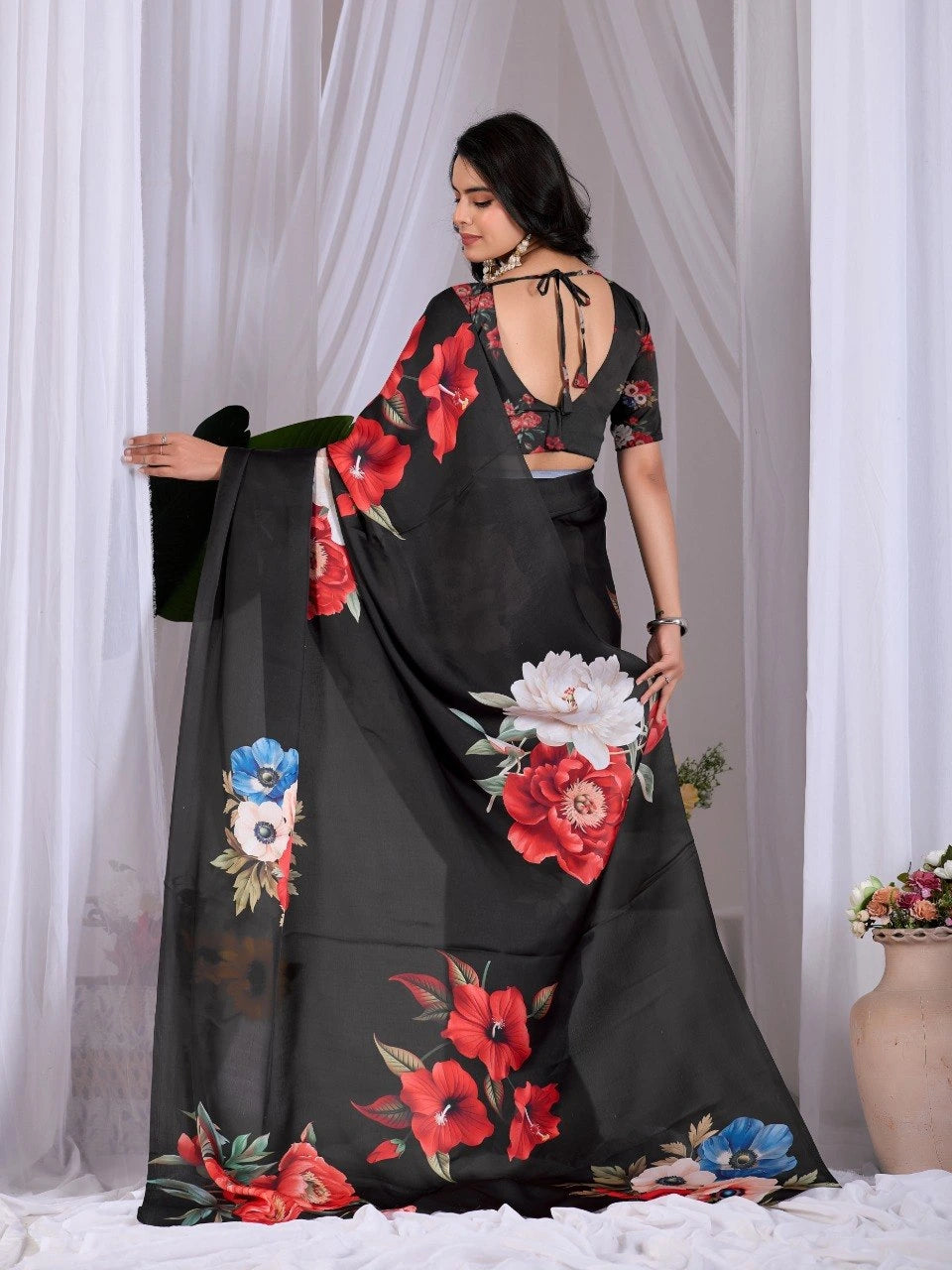 Woman wearing a black saree with floral patterns in front of white curtains