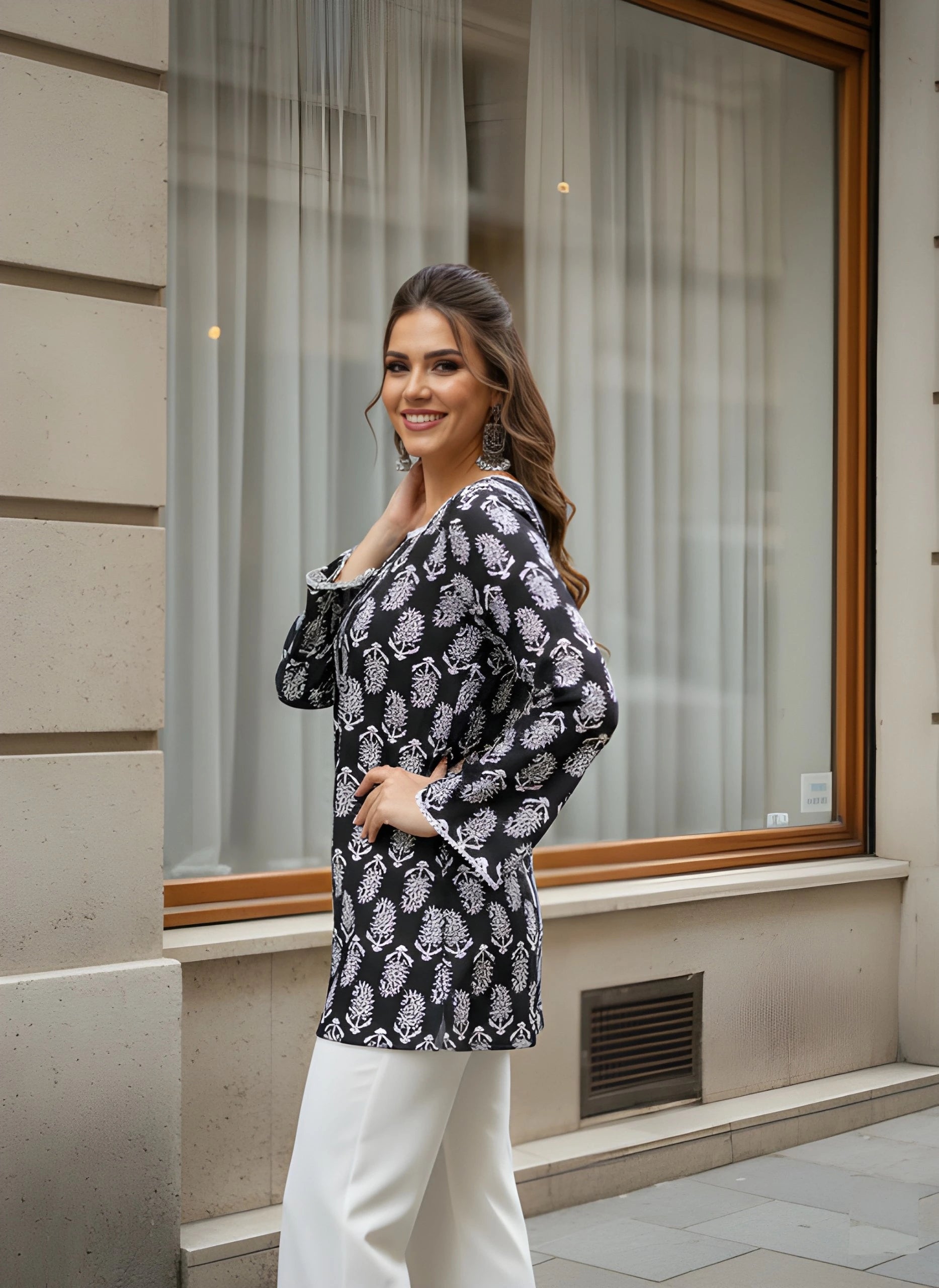 Woman in a black and white patterned blouse and white pants standing in front of a store window.