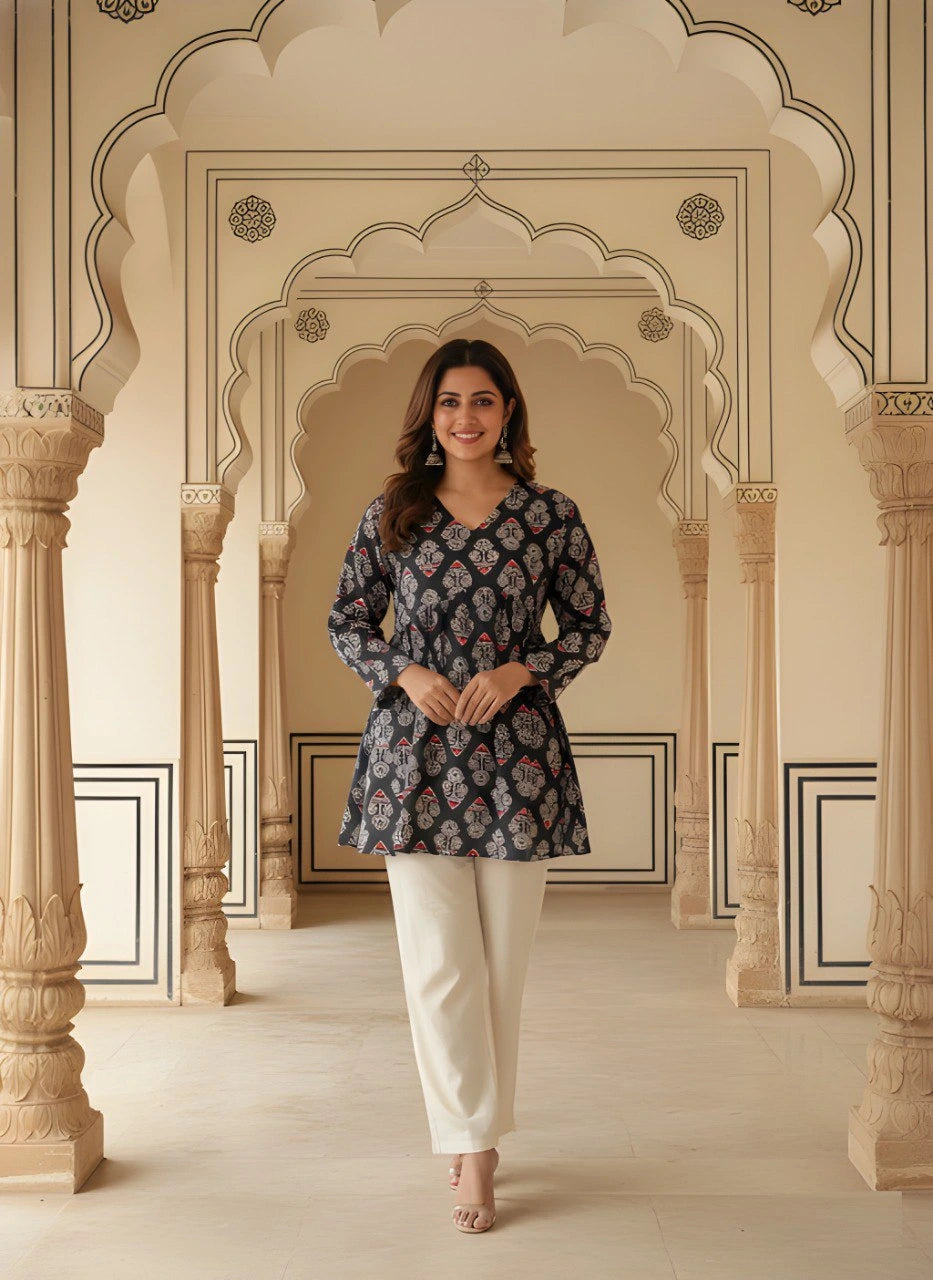 A woman stands facing forward in a black short kurti featuring a white and red ethnic block print. The top has a V-neckline, long sleeves, and a gathered empire waist that flares into a peplum hem. She is wearing off-white trousers and large silver jhumka earrings.