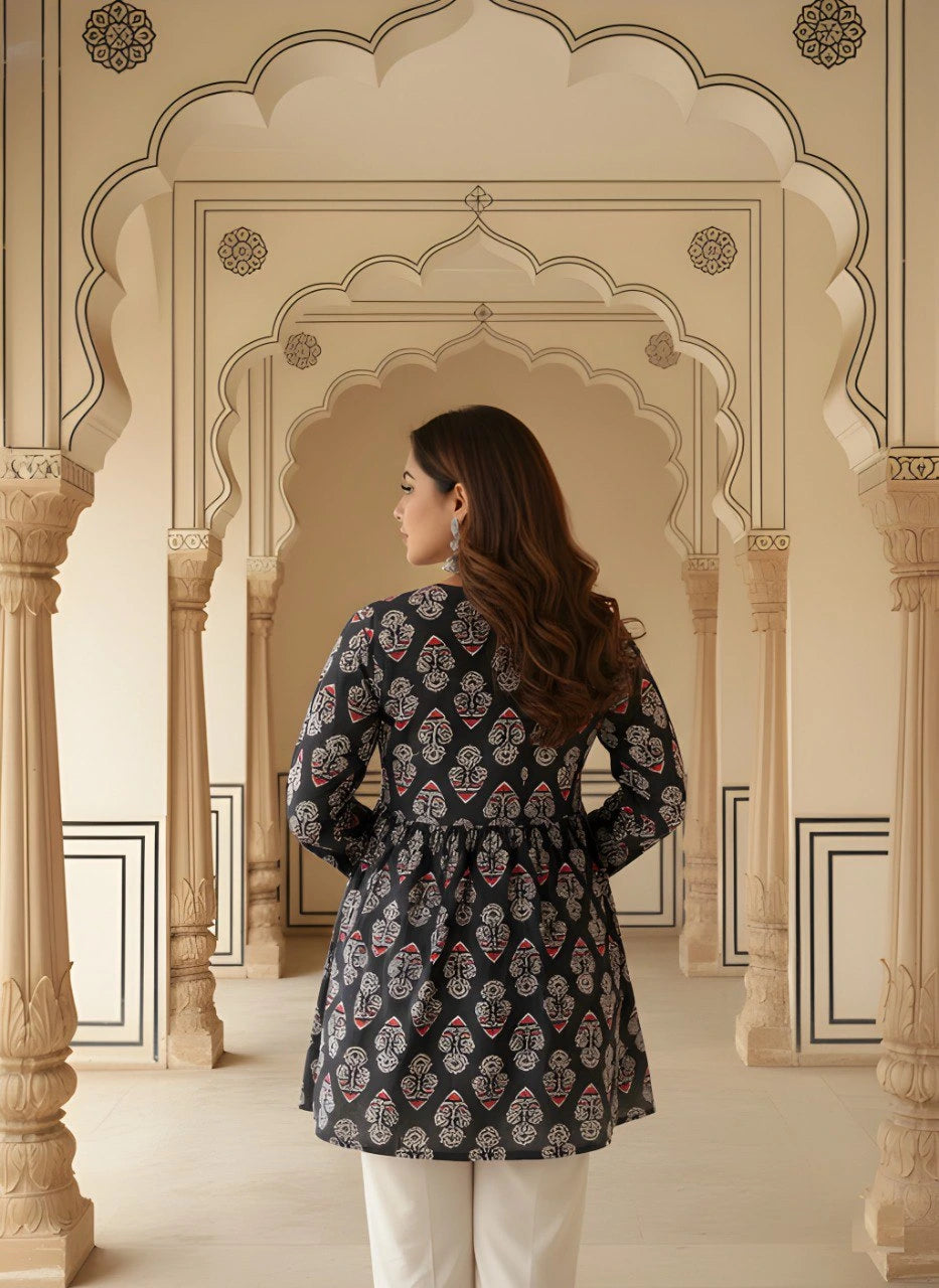 A rear view of the woman showcasing the gathered waistline and the continuous block print across the back of the kurti. She is standing between ornate pillars in a traditional architectural hallway.