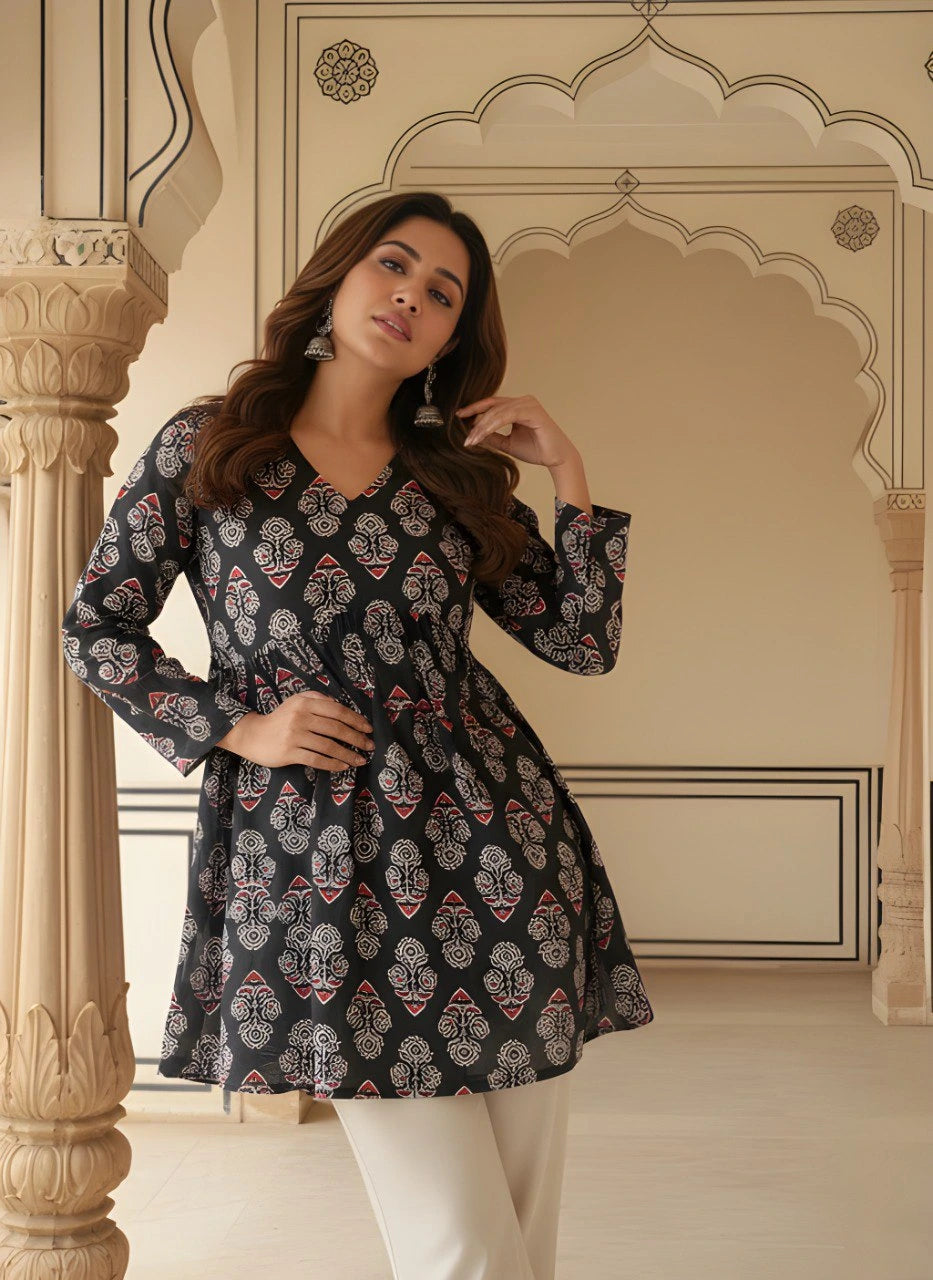 A wide shot of the woman walking toward the camera in a palace-like corridor. The full ensemble—the printed kurti, white straight-fit trousers, and silver accessories—is visible against the neutral-toned, symmetrical background.