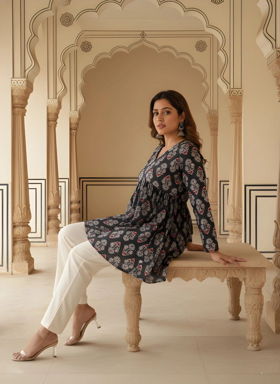 The woman is seated on a carved stone bench, leaning back slightly. This angle highlights the fluid drape of the peplum flare and the intricate details of the floral and geometric motifs. She pairs the look with clear-strapped heels.
