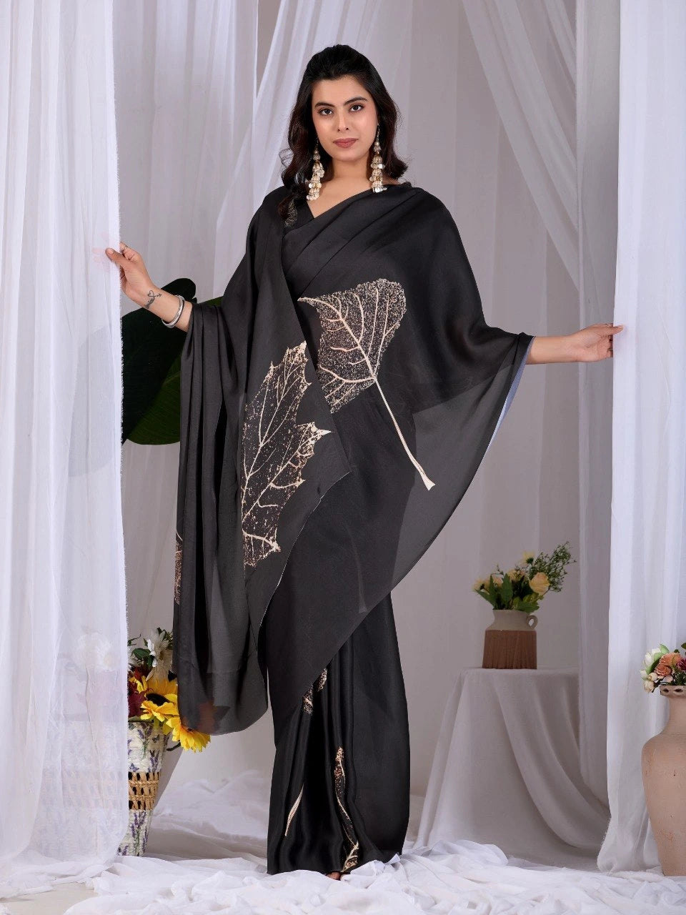 Woman wearing a black saree with leaf patterns in front of white curtains.