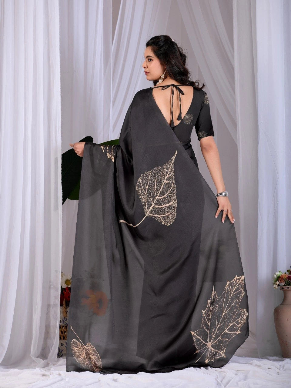 Woman wearing a black saree with leaf patterns against a white curtain background