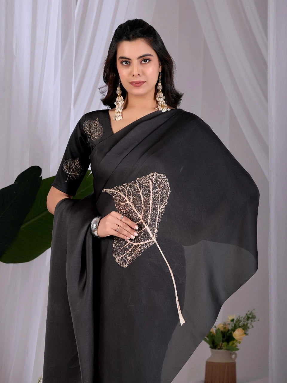 Woman wearing a black saree with leaf design against a white curtain background