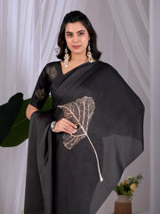 Woman wearing a black saree with leaf design against a white curtain background