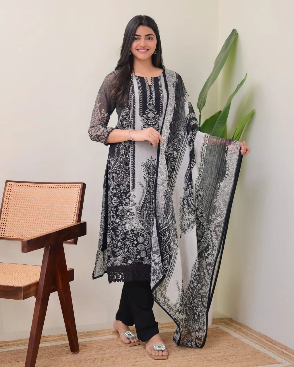 Woman holding a black and white patterned dupatta in a room with a chair and plant.