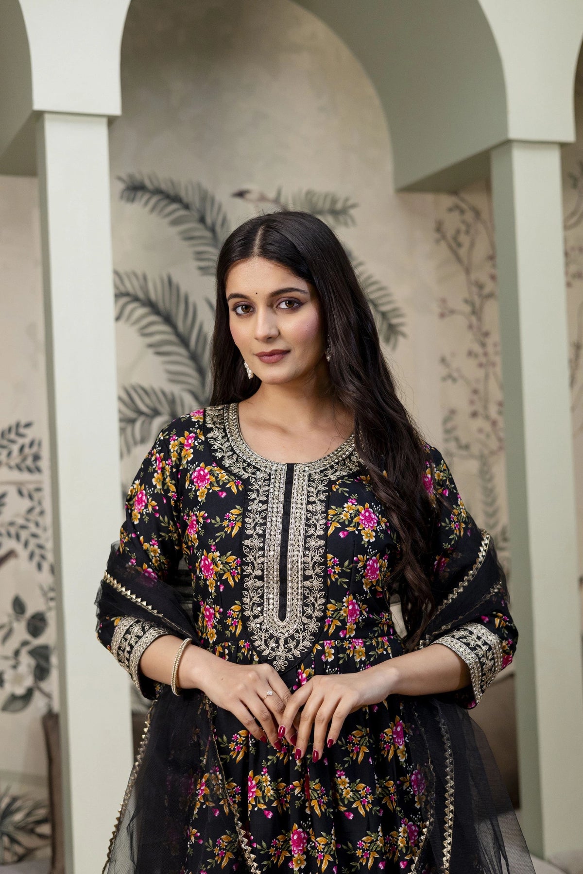Woman wearing a black embroidered traditional outfit with floral patterns in an indoor setting.
