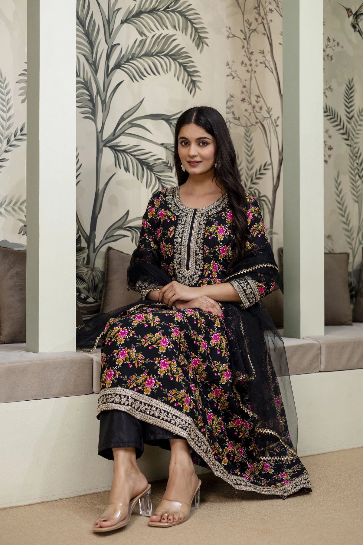 Woman in a floral dress sitting in a decorated room with plants.