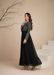 Woman in a black and patterned long dress standing on a staircase with a beige wall and branch in the background.