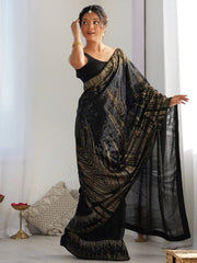 Woman wearing a black and gold saree in a room with white curtains and a pillow.