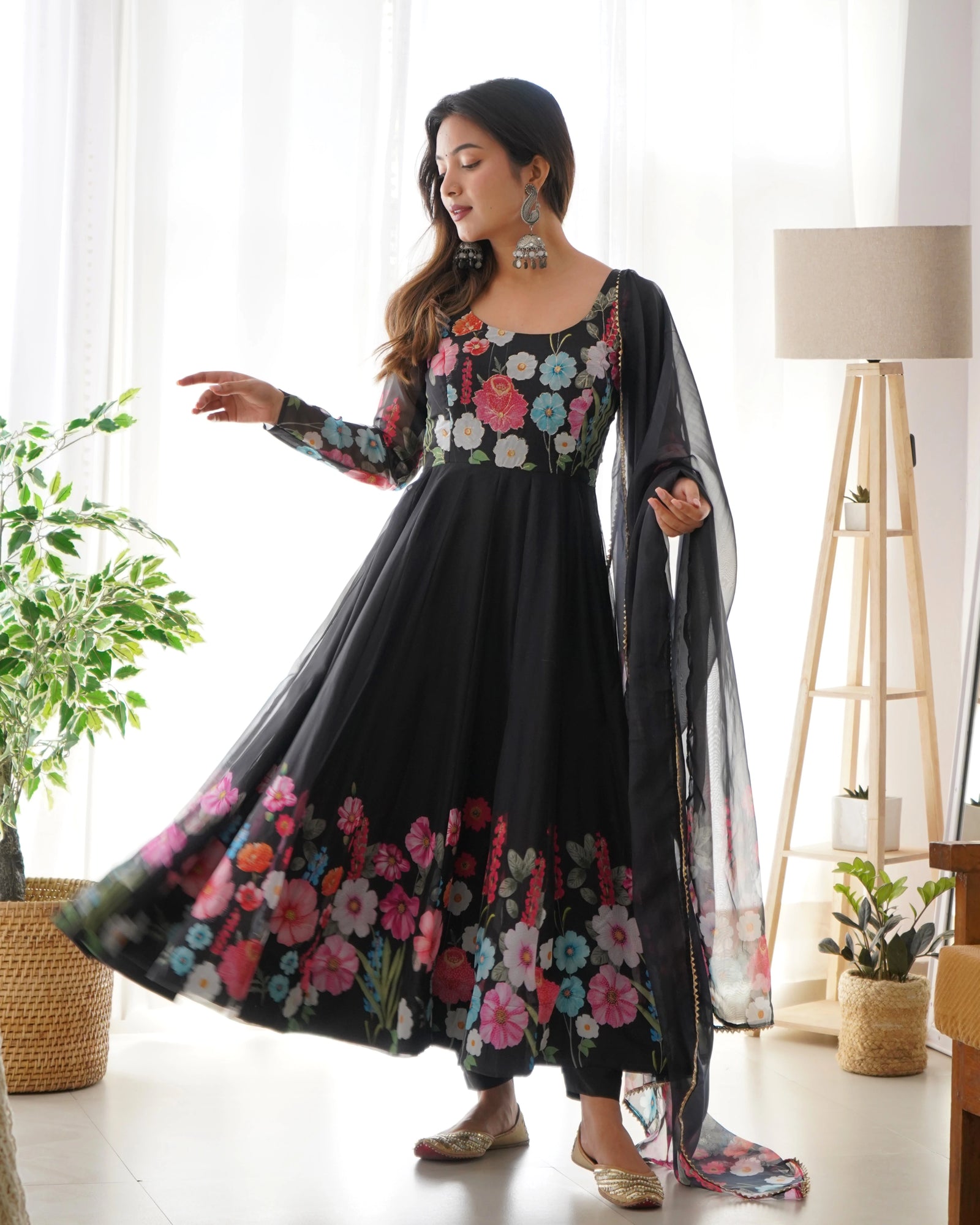 Woman wearing a black floral dress in a bright room with plants and a lamp.