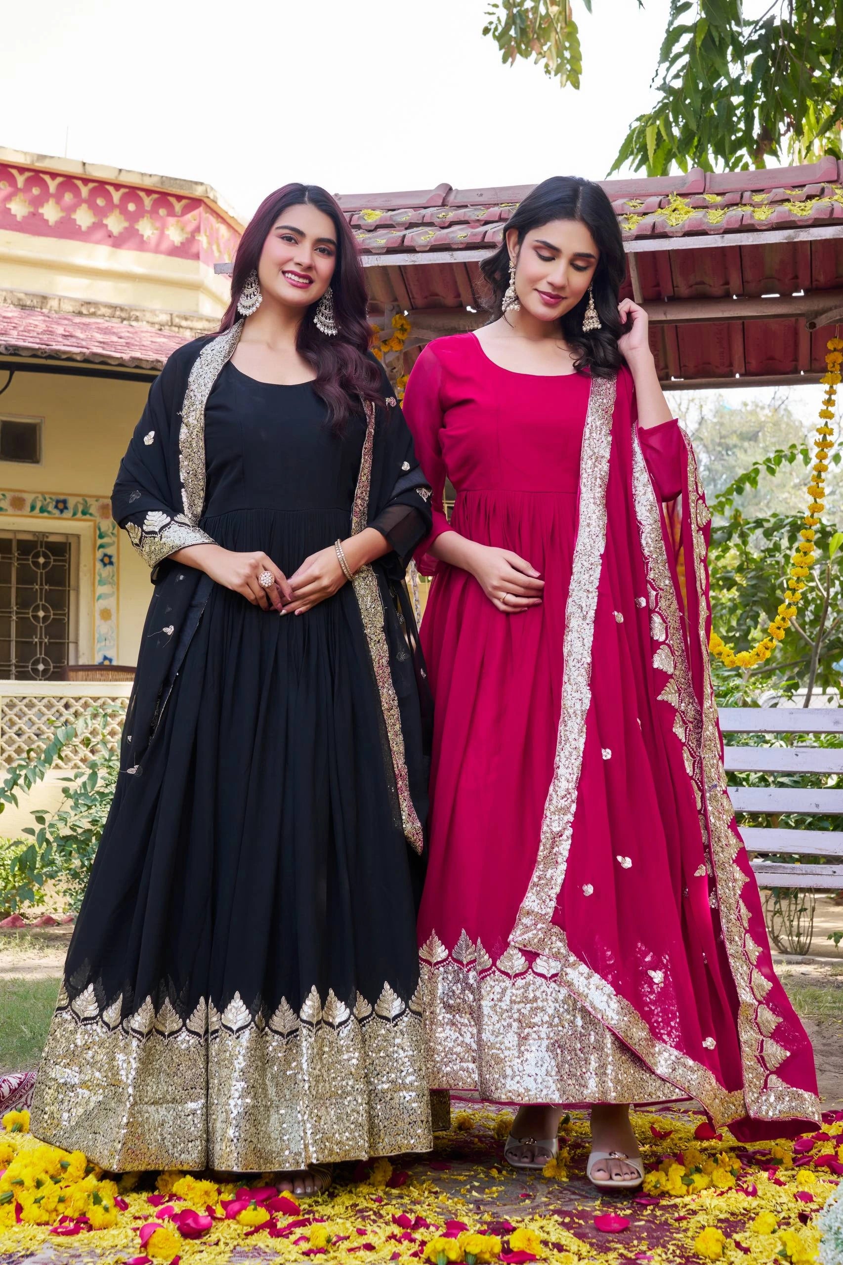 Two women in traditional black and pink dresses standing outdoors with a decorative background.