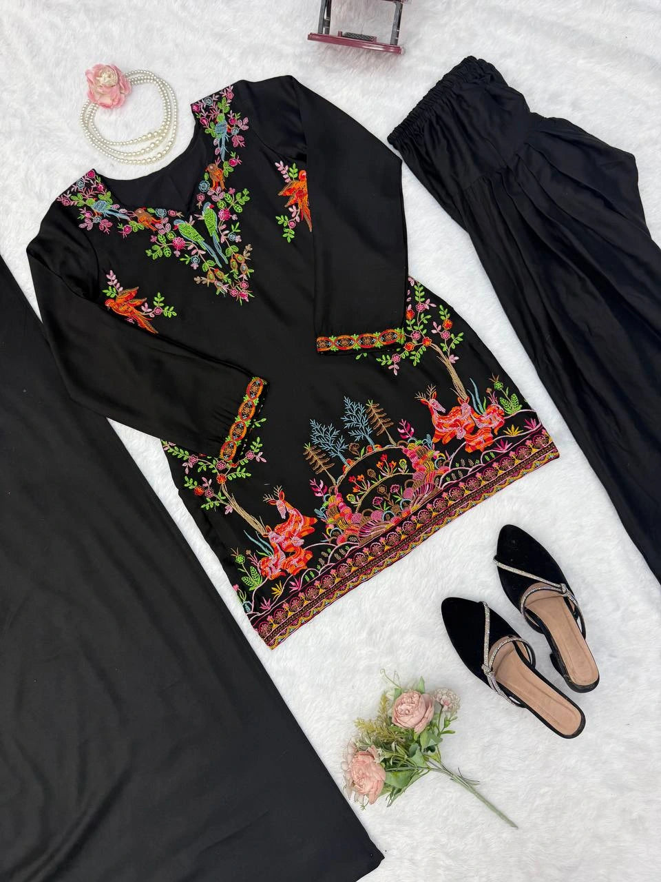 Black outfit with colorful embroidery on a white surface with shoes and flowers.