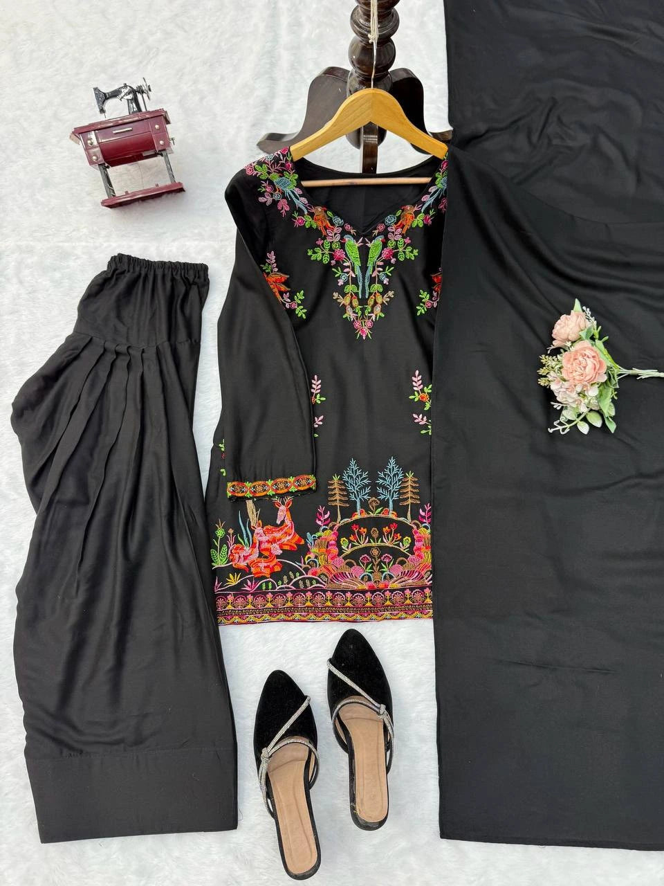 Black embroidered dress with floral patterns, a hanger, and a small bag on a textured white background.
