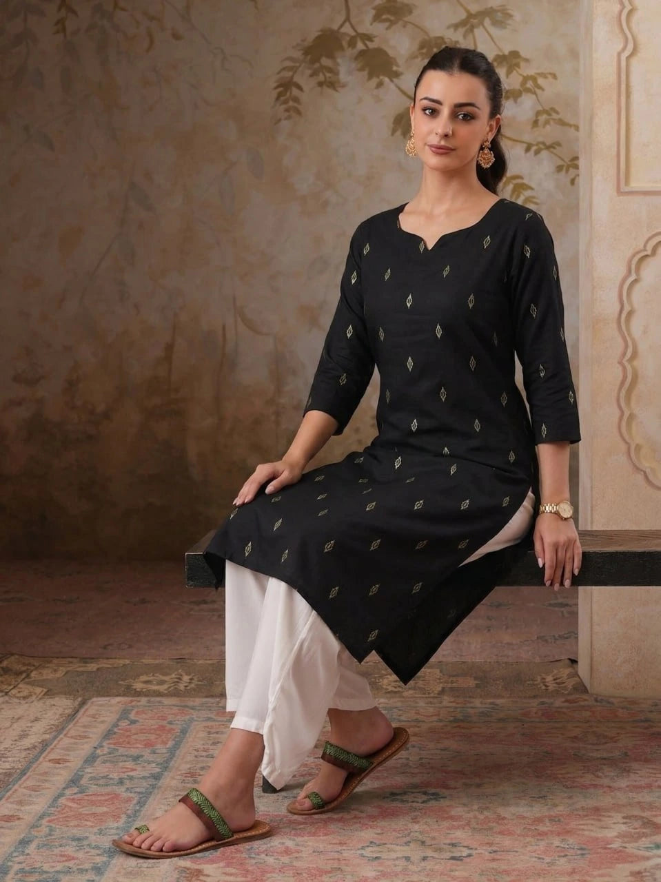 Woman wearing a black kurta with white pants sitting on a decorative rug.