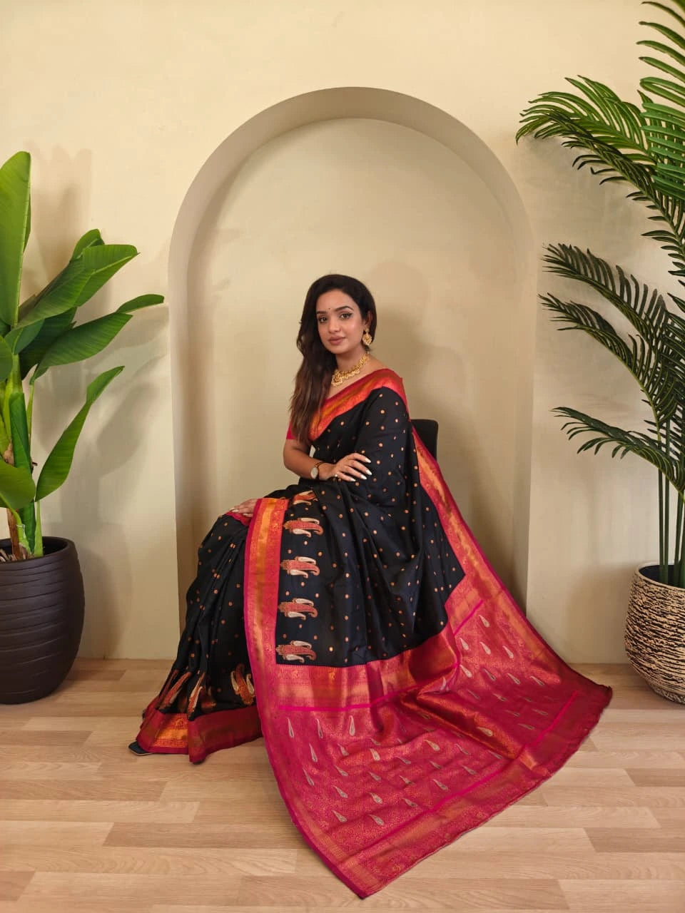 Woman in a black and red saree sitting in a room with plants.