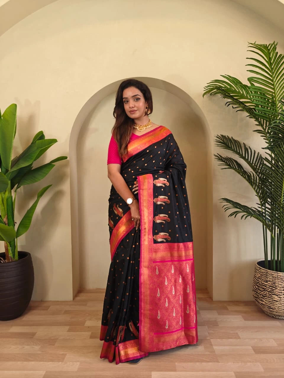 Woman in a black and pink saree standing in a room with plants.