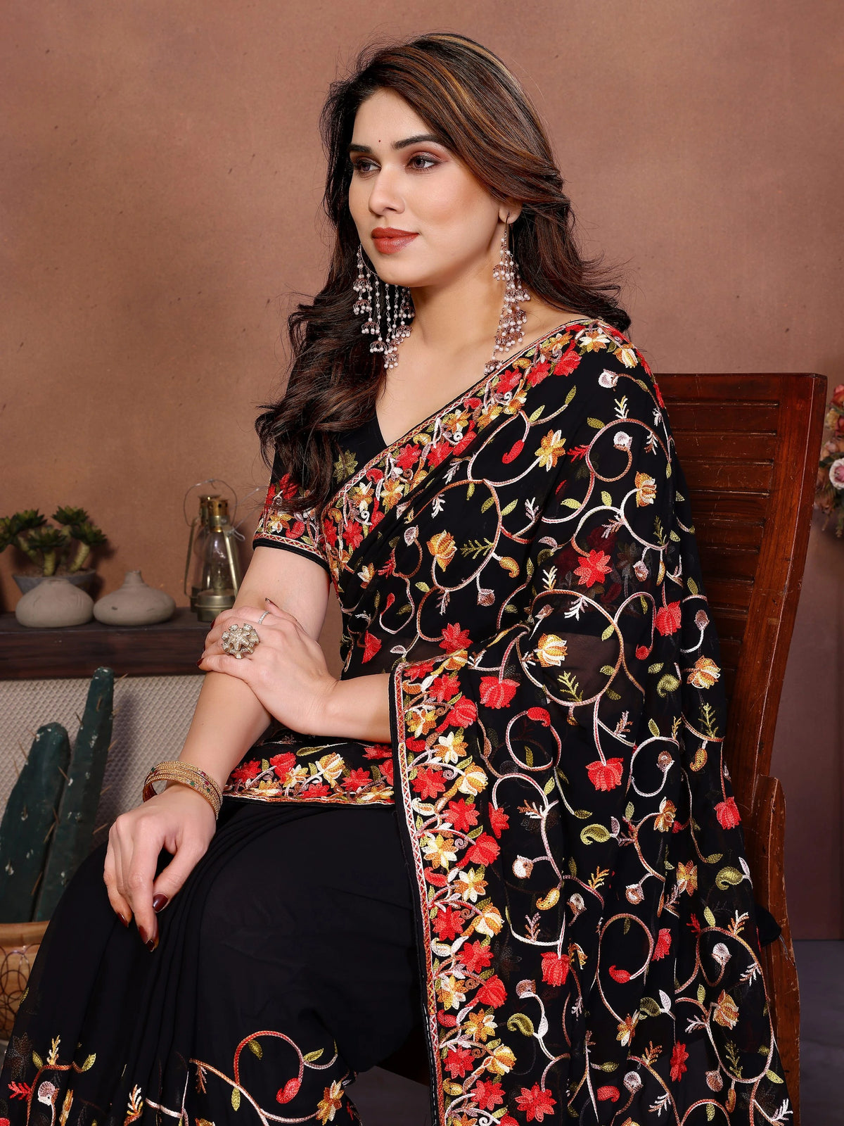 Woman wearing a black embroidered saree with red and gold details, sitting against a brown background.
