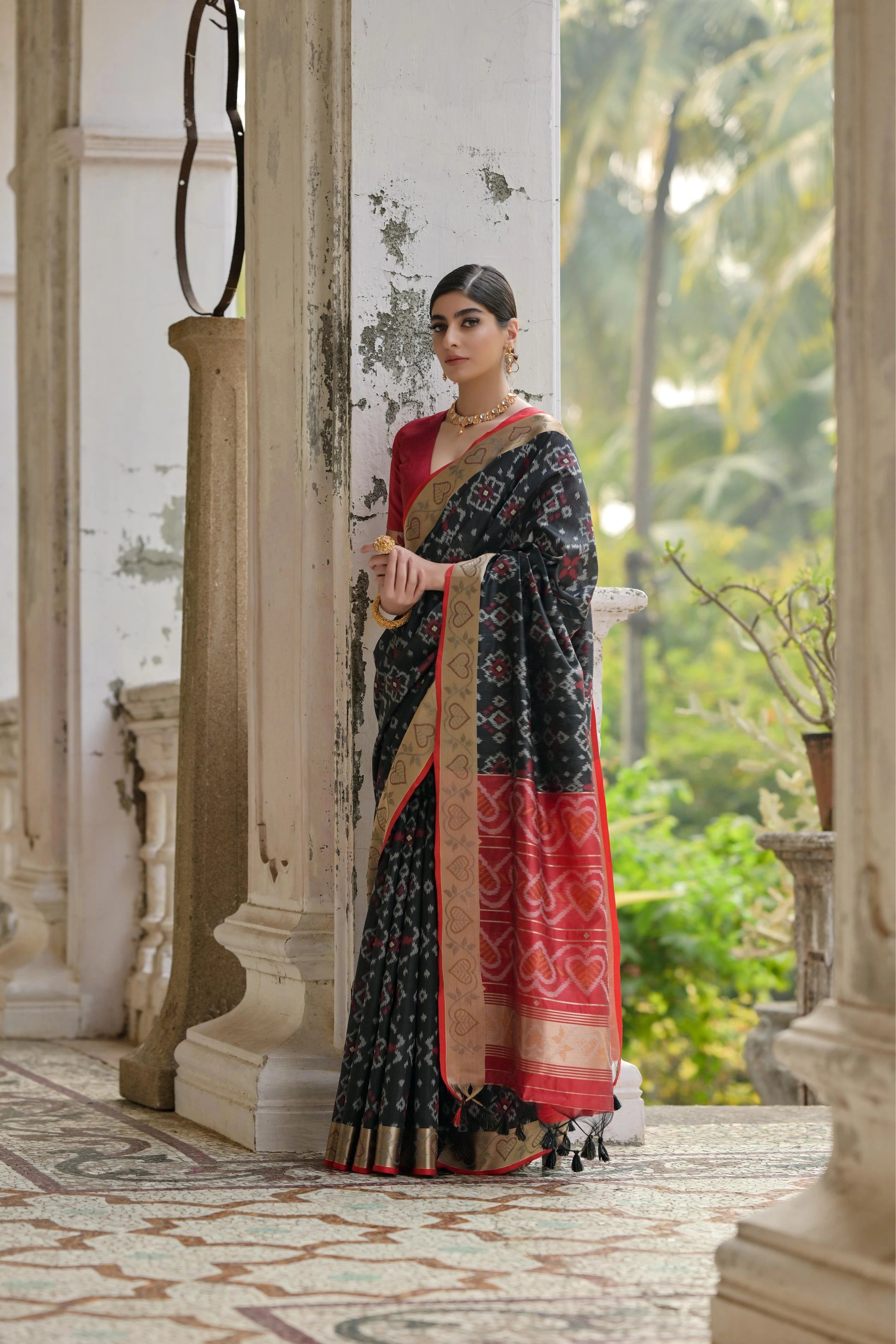Woman in a traditional saree standing in an elegant outdoor setting with columns and greenery.
