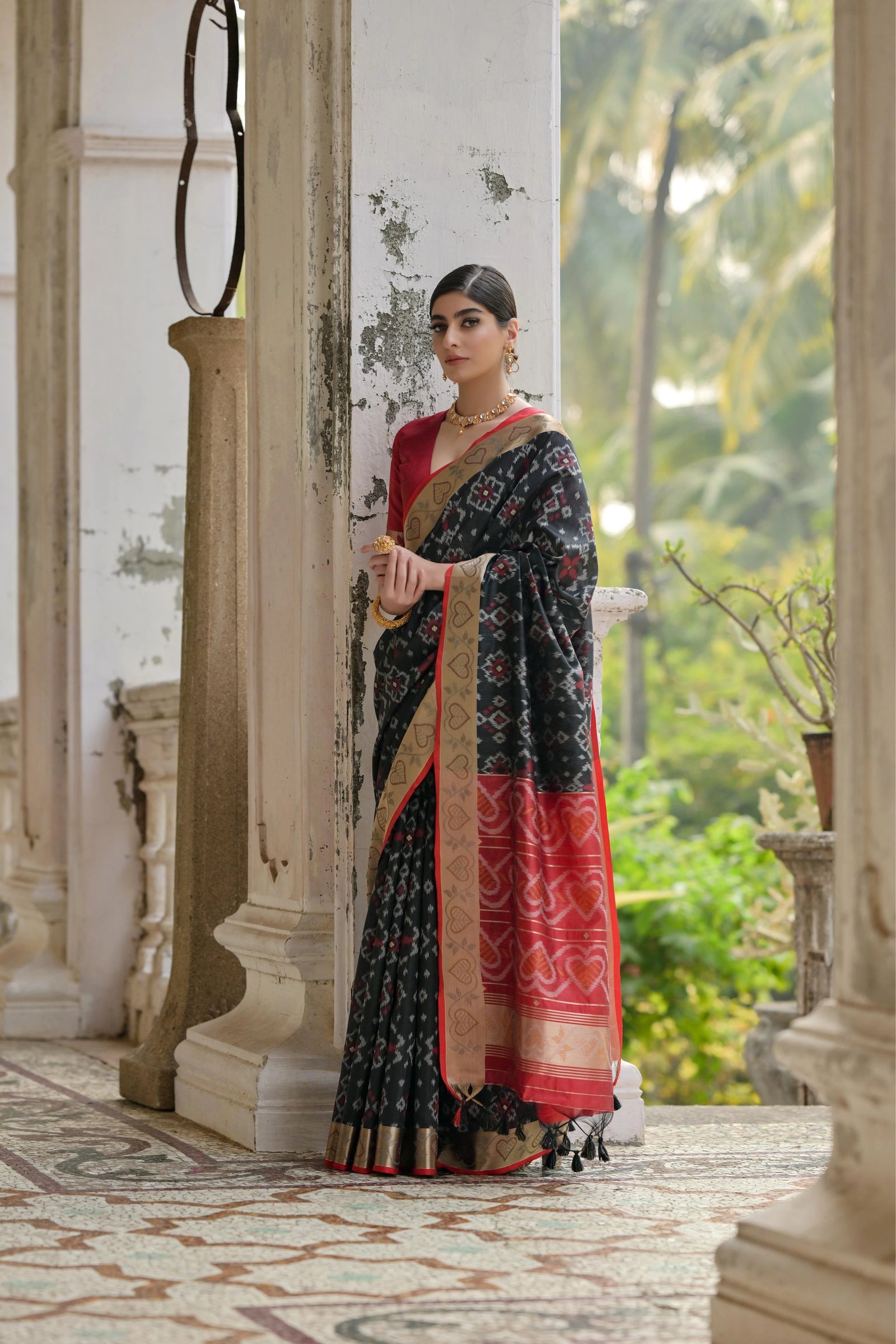 Woman in a traditional saree standing in an elegant outdoor setting with columns and greenery.
