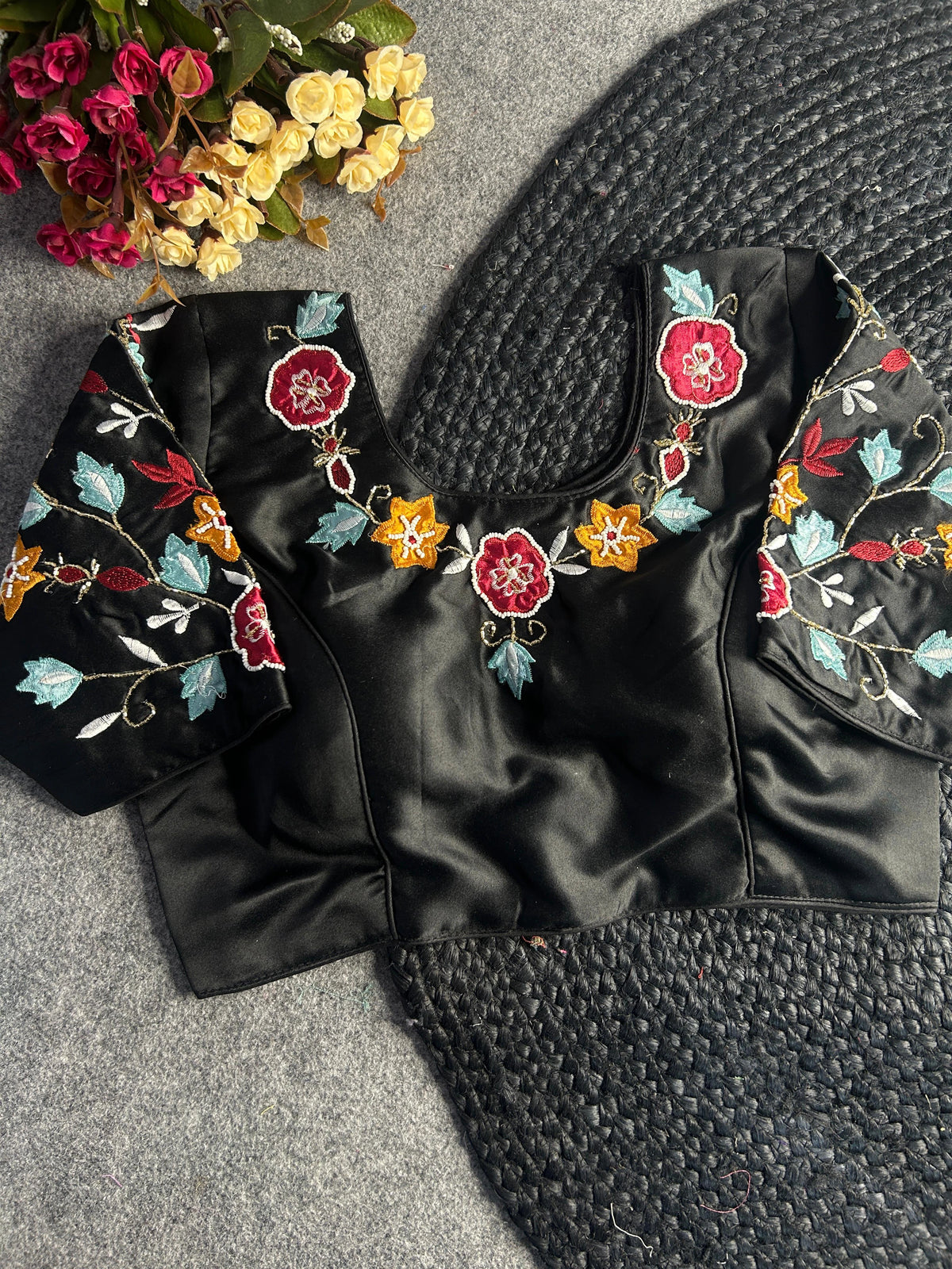 Back view of the same black blouse, featuring a centered hook-and-eye closure. The floral embroidery continues along the back neckline and across the sleeves, set against the smooth, lustrous finish of the black fabric.