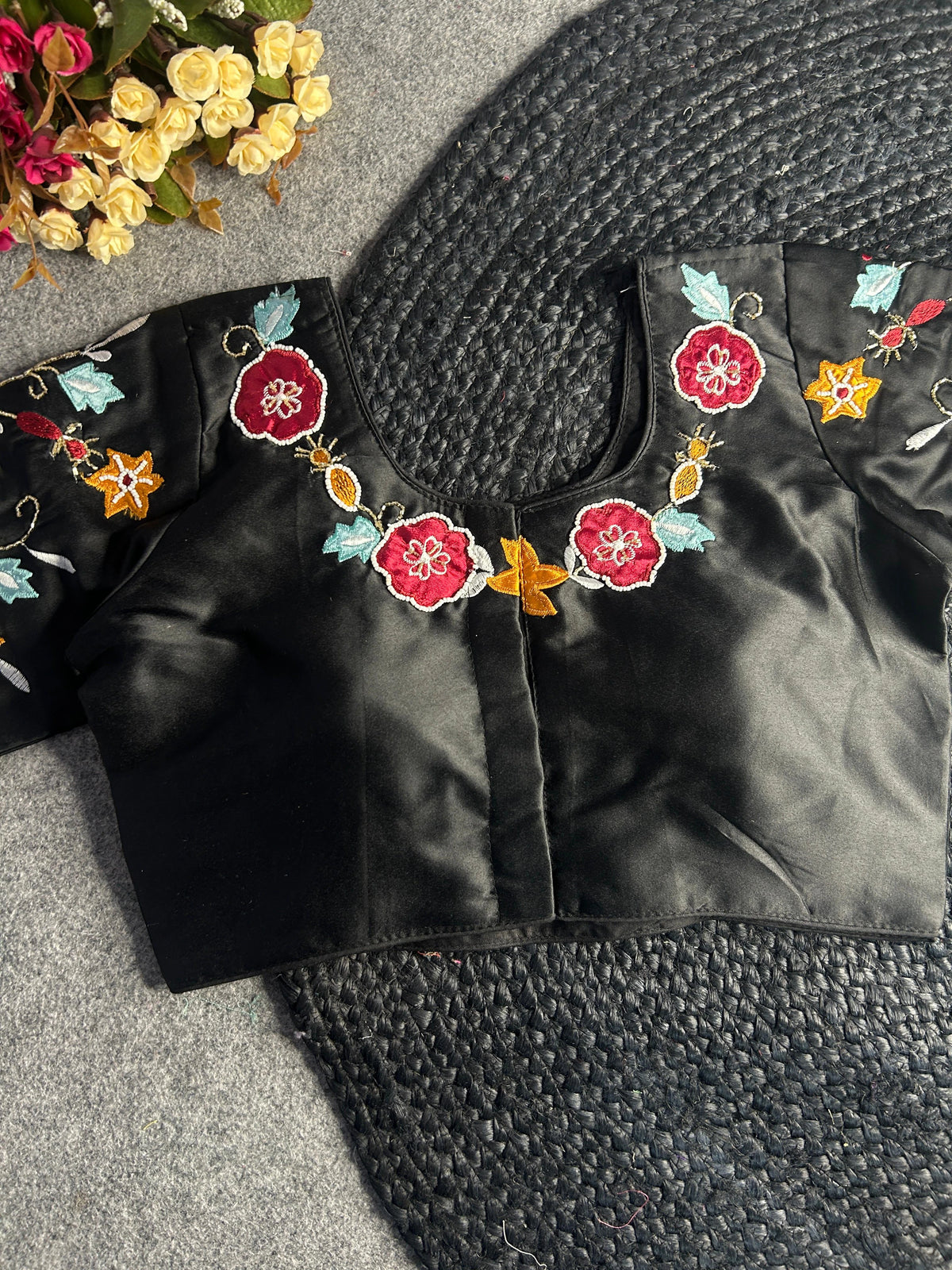 Front view of a black silk-satin blouse with a wide U-neckline. The neckline and sleeves are decorated with vibrant floral embroidery in shades of ruby red, mustard yellow, and light blue, accented with delicate gold vine stitching.