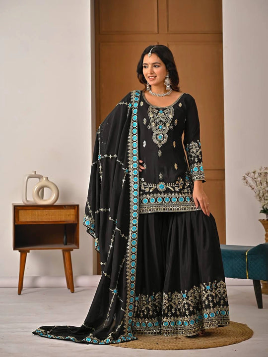 Woman wearing a black embroidered traditional outfit with turquoise details in a room setting.