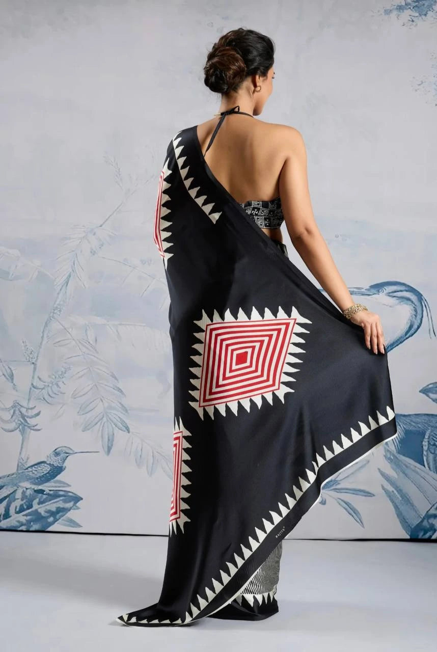 Woman wearing a black saree with red and white patterns, standing against a light gray background.