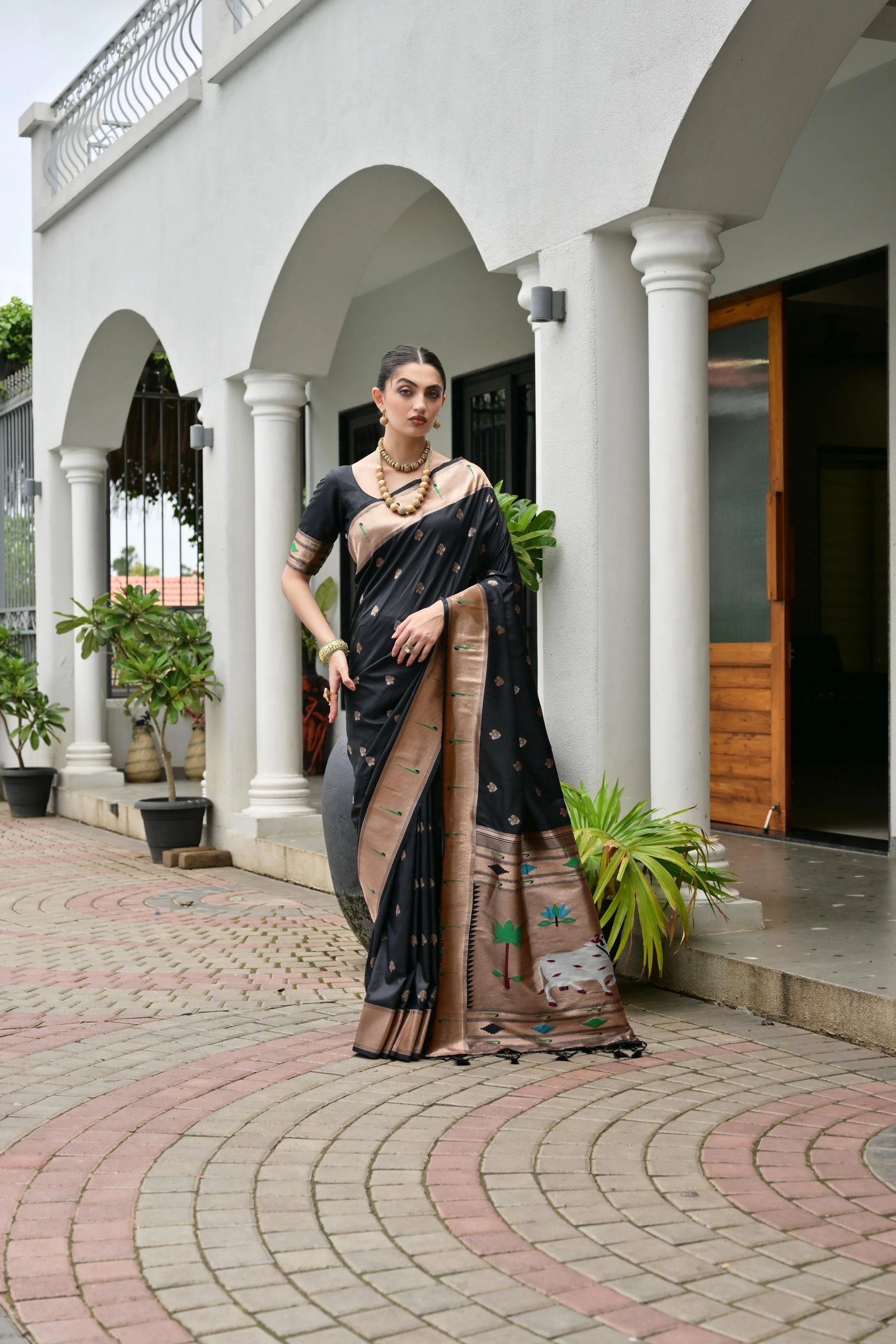 Woman in a black and brown saree standing in front of a white building with columns.