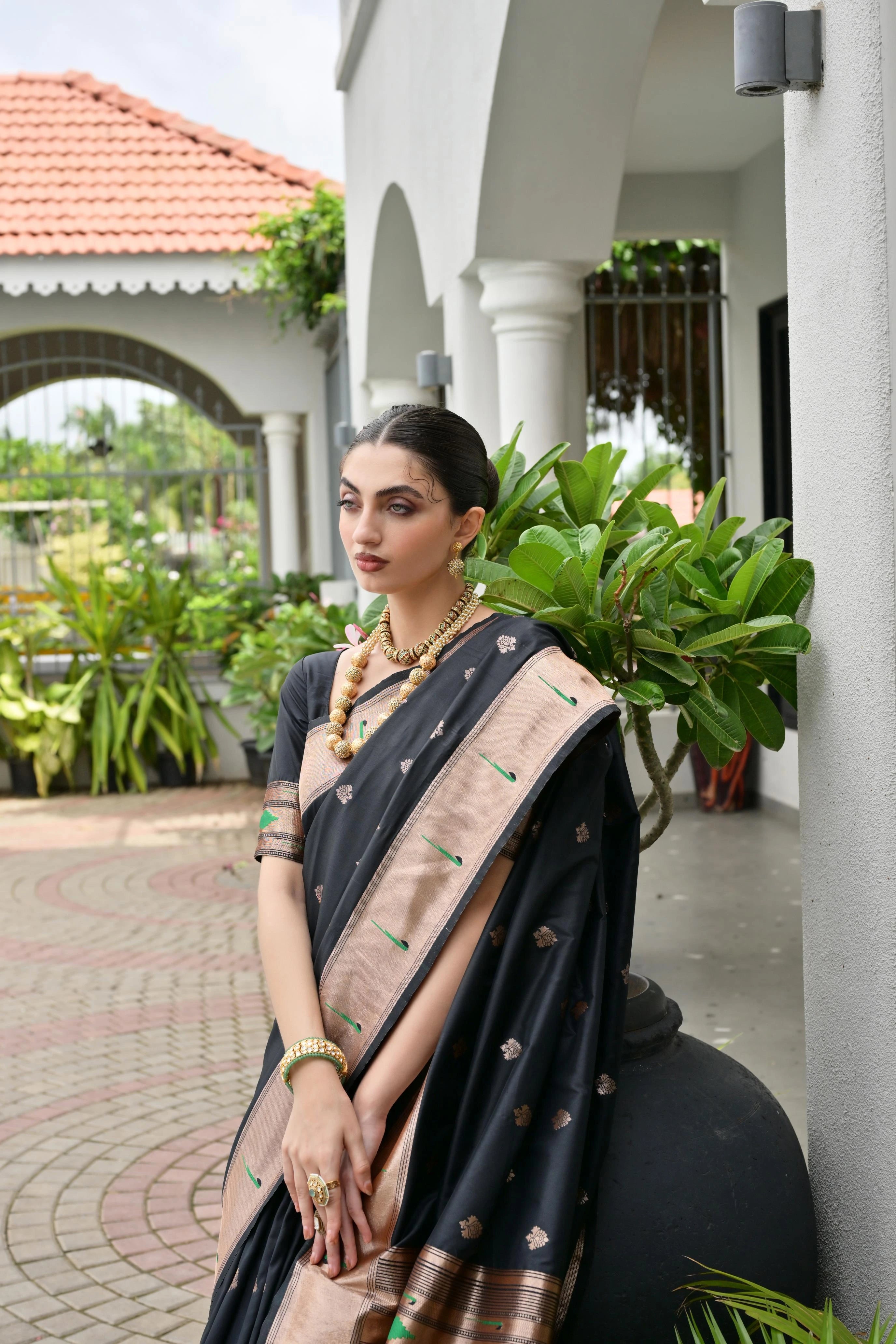 Woman in a black and beige saree standing outdoors with greenery and a building in the background