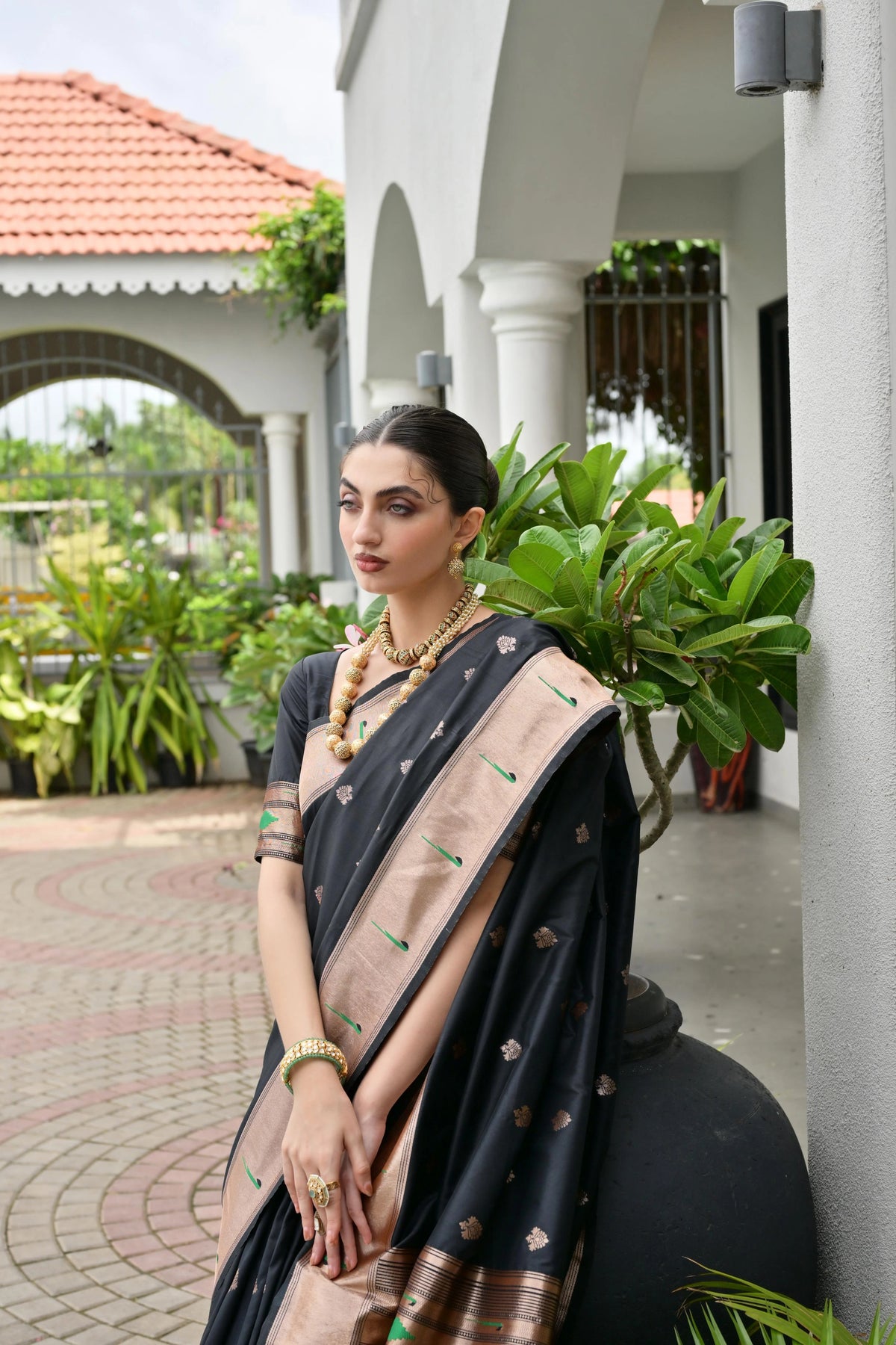 Woman in a black and beige saree standing outdoors with greenery and a building in the background
