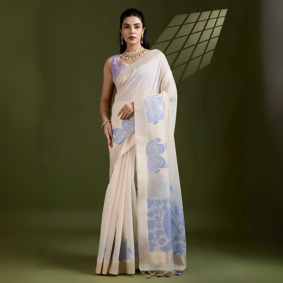 A woman models an elegant Beige Soft Muga Cotton Saree. The saree features large Kalamkari-style prints of peacocks and florals in a contrasting Soft Lavender Blue on the pallu. She wears a sleeveless lavender blouse and accessorizes with a gold and pearl Kundan choker necklace, posing indoors.