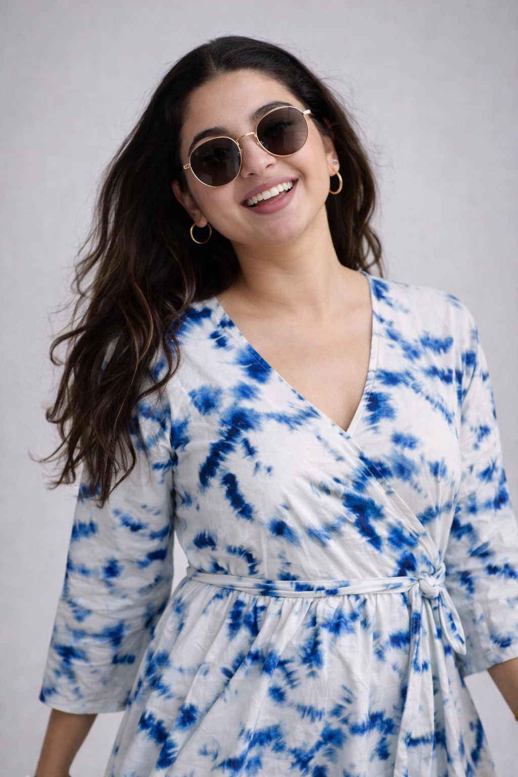 Woman wearing a blue and white tie-dye dress with sunglasses against a gray background